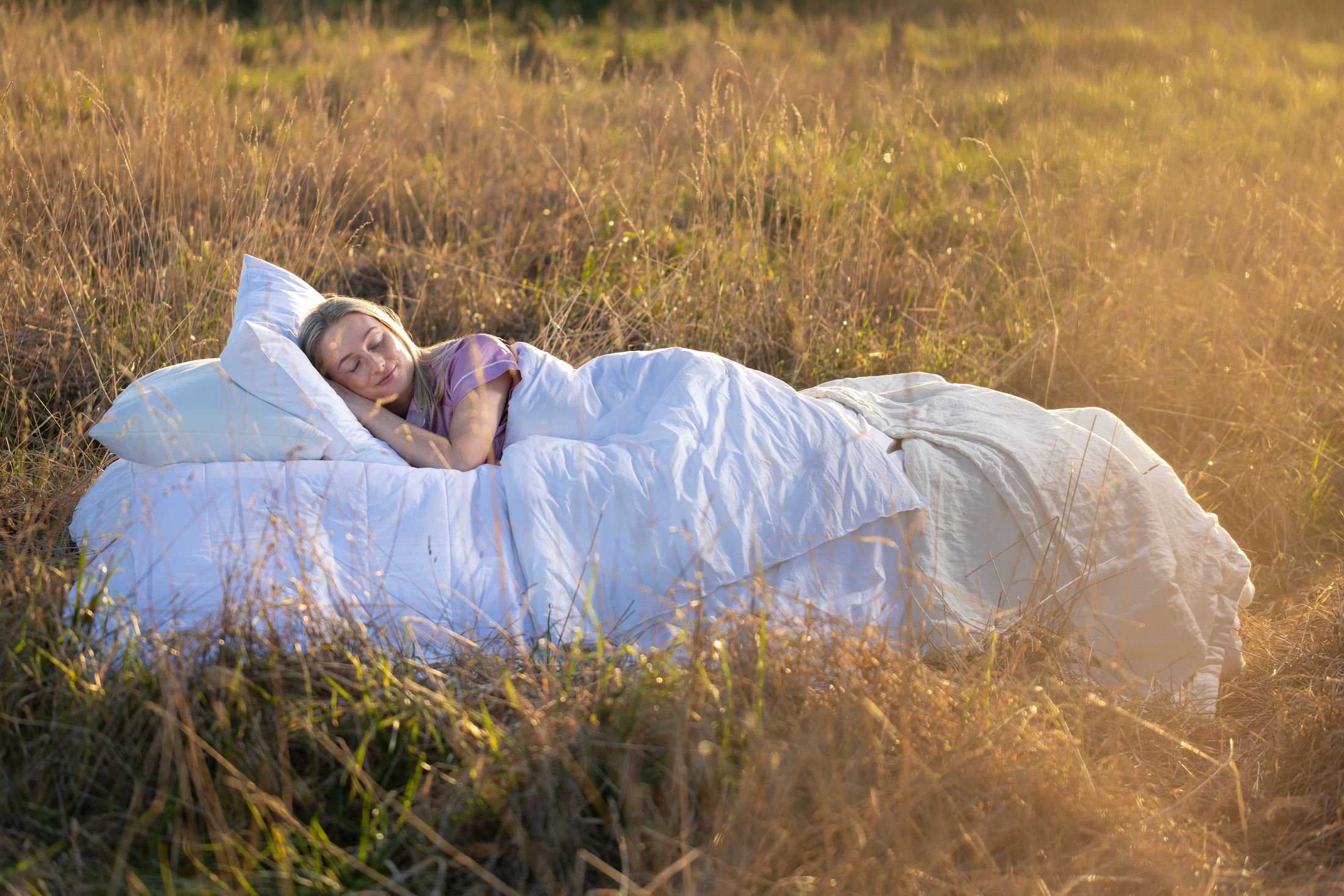 A woman sleeping on a white blanket in a grassy field during sunset.