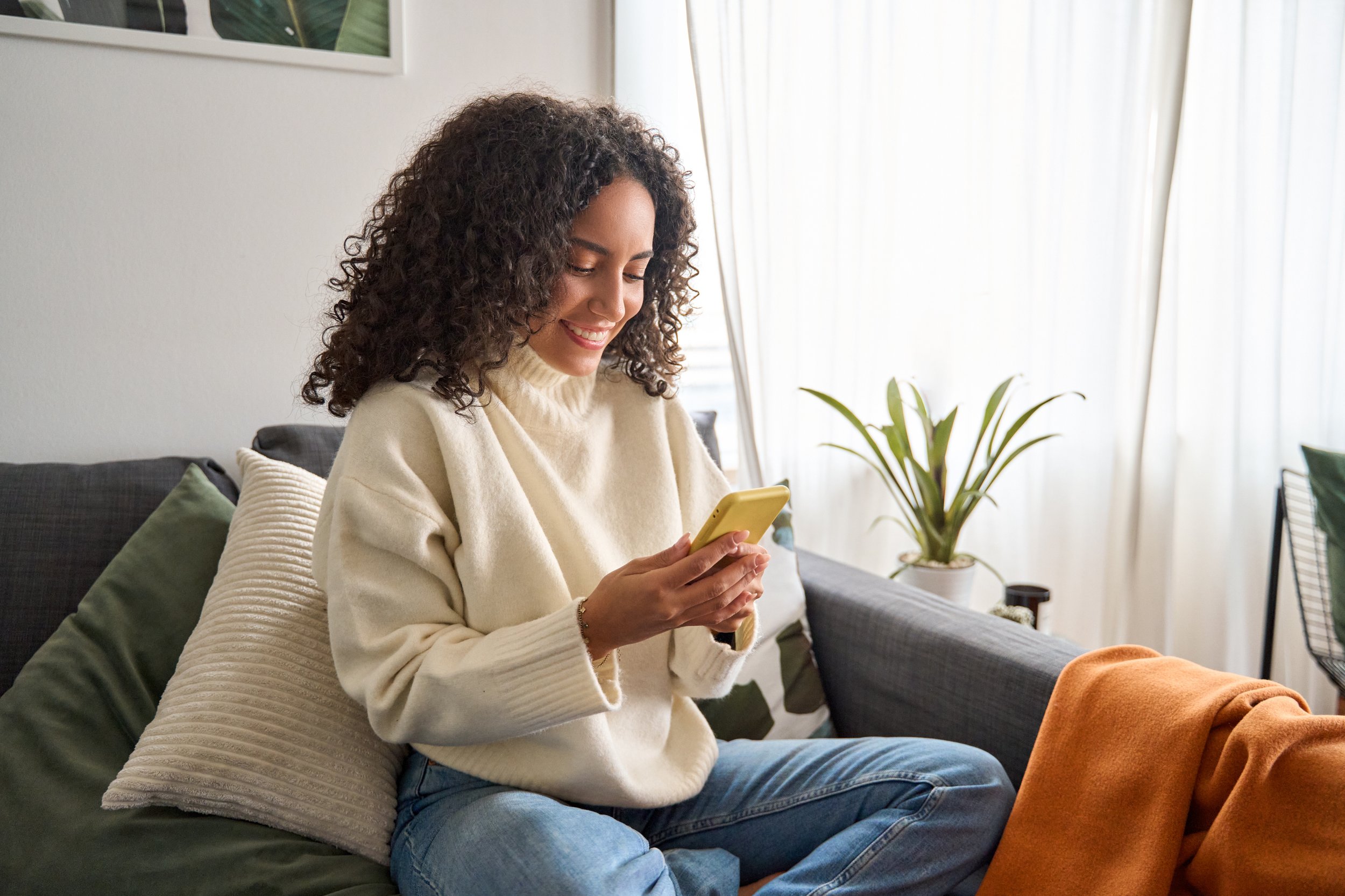 A woman in a yellow sweater looking at her phone smiling appearing to be on a Virtual Care call with a doctor in her living room.