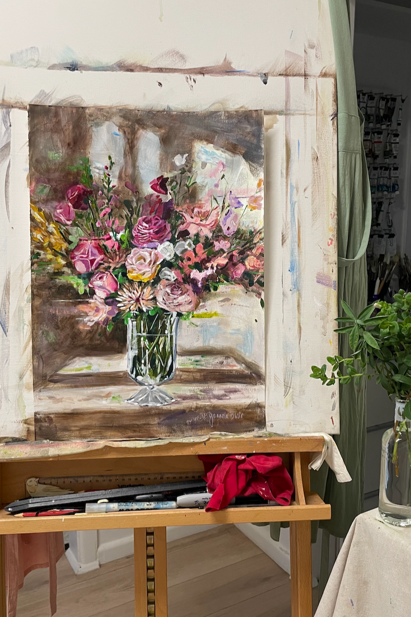 Oil painting of a lush pink floral bouquet in a glass vase, displayed on an easel in the artist’s studio with brushes, cloths, and greenery nearby.