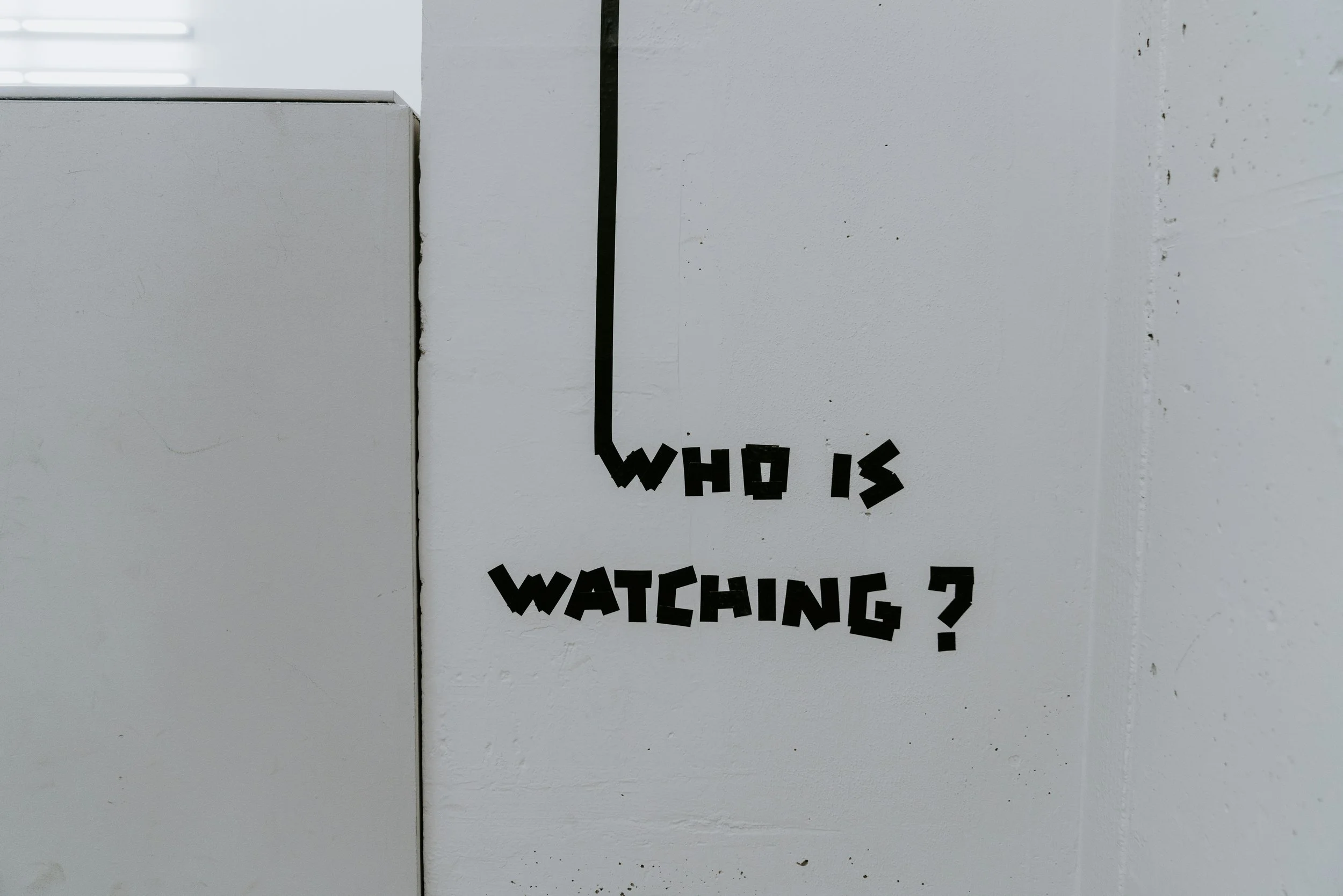 Who is watching written on a wall under a cable that might lead to a security camera