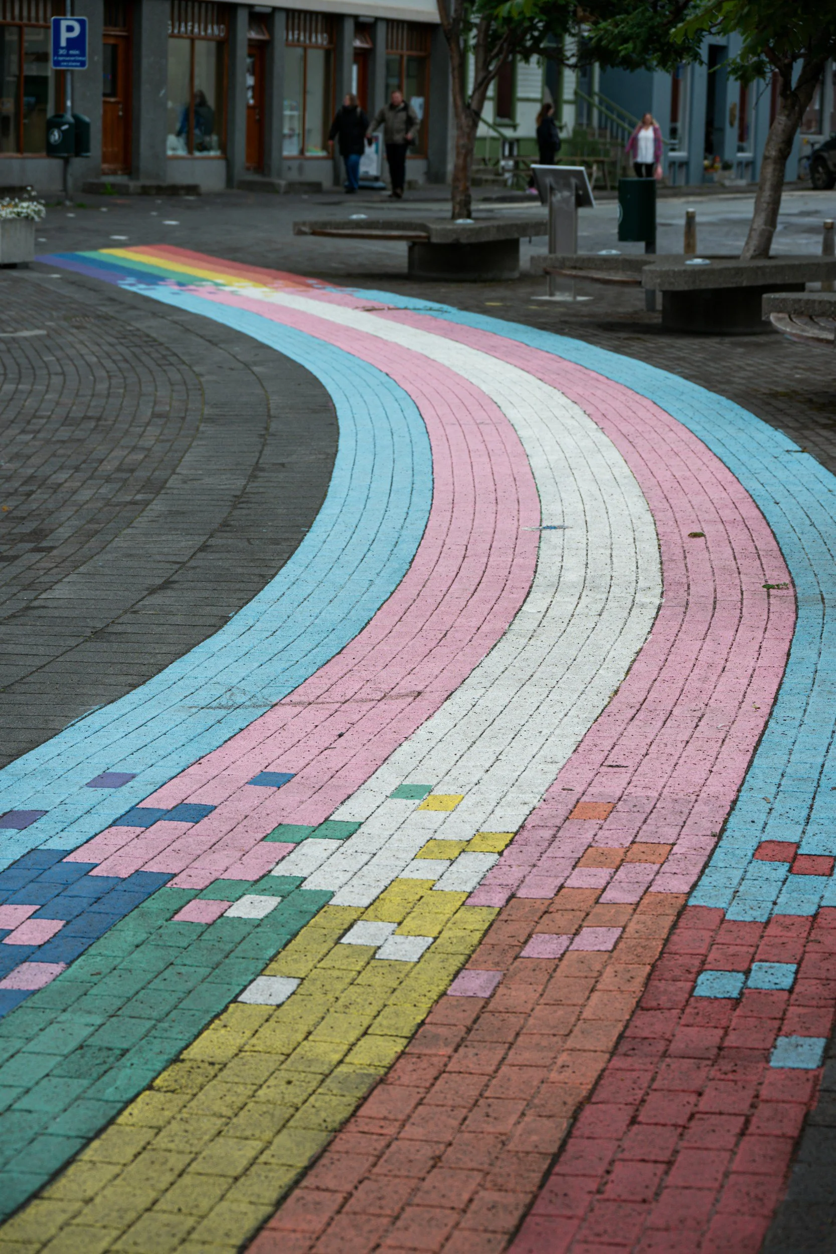 A brick sidewalk painted with a colorful rainbow design, crossing a small street in an urban area with shops and people walking.