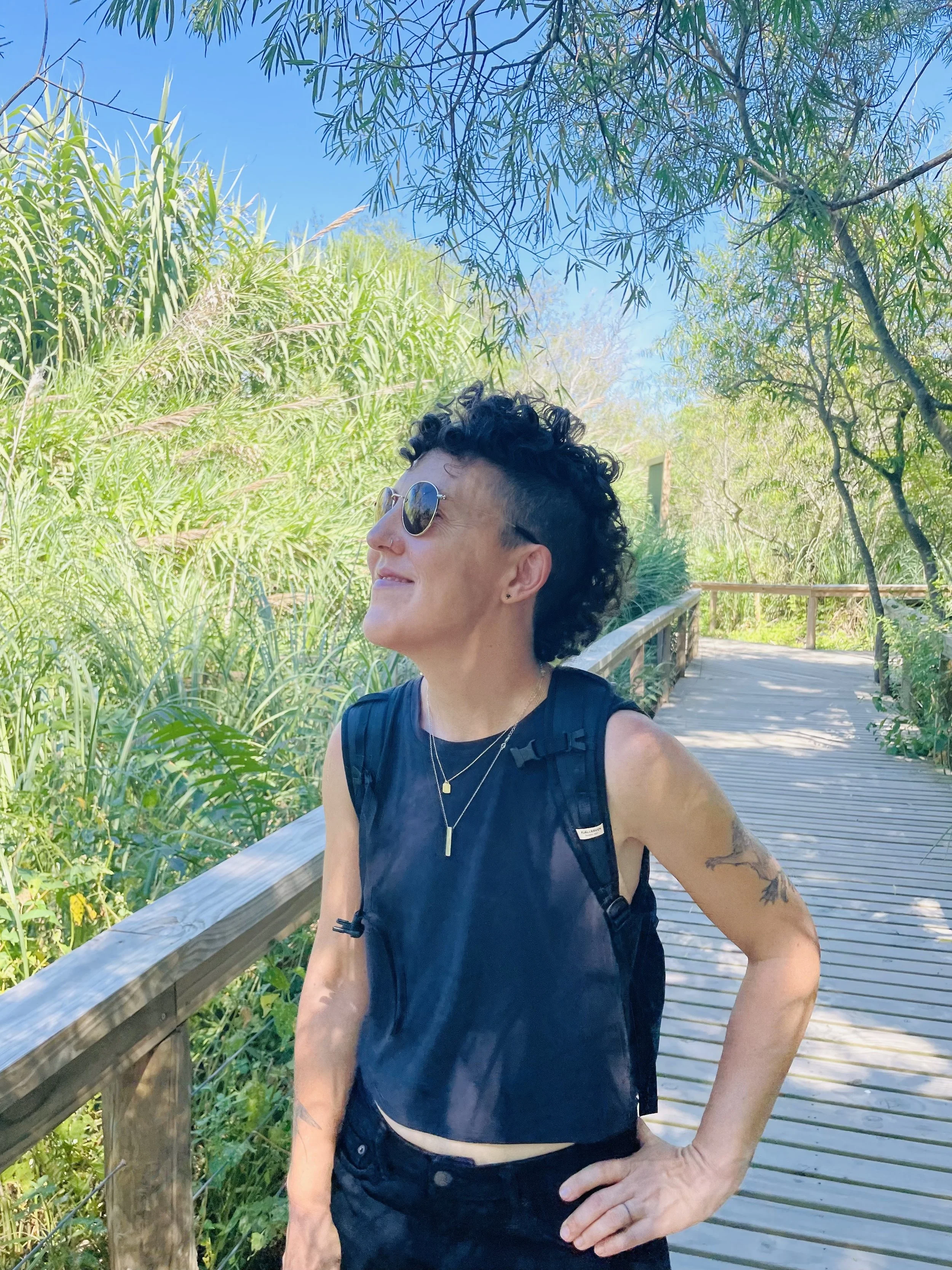 A woman with short curly black hair and sunglasses standing on a wooden walkway surrounded by lush green plants and trees under a clear blue sky.