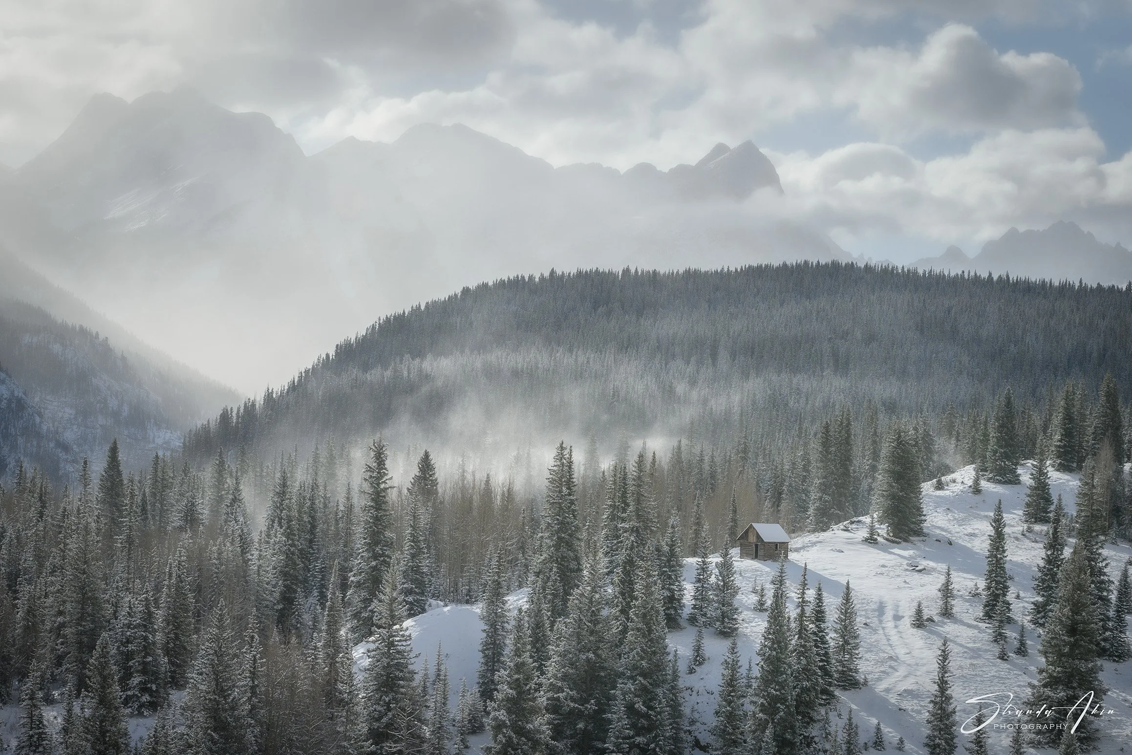 Snow-covered mountain landscape with dense pine forest and a small cabin in the foreground. Fog and clouds are present in the sky.
