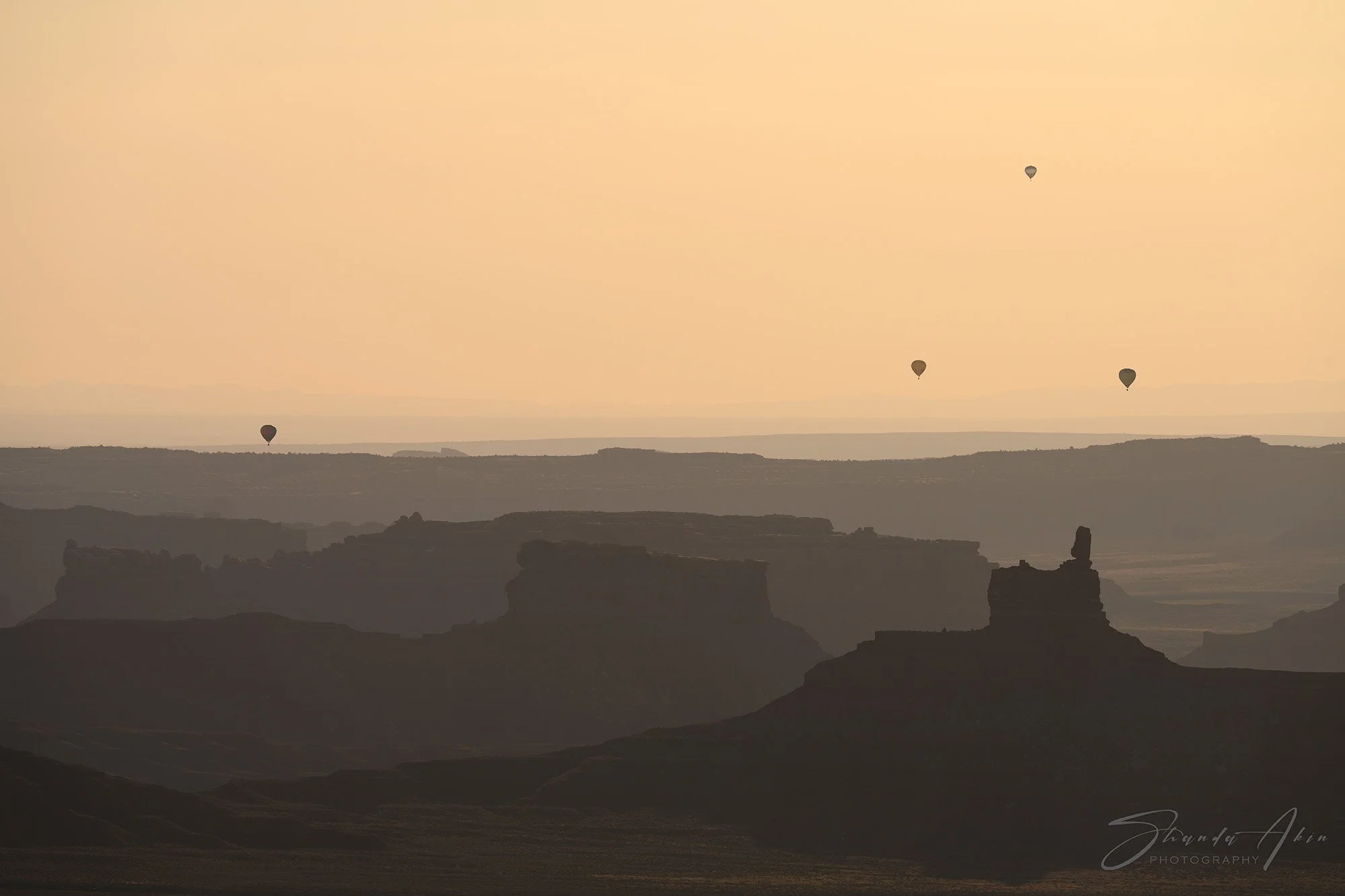 A desert landscape with layered rock formations and four hot air balloons in the sky during sunset.
