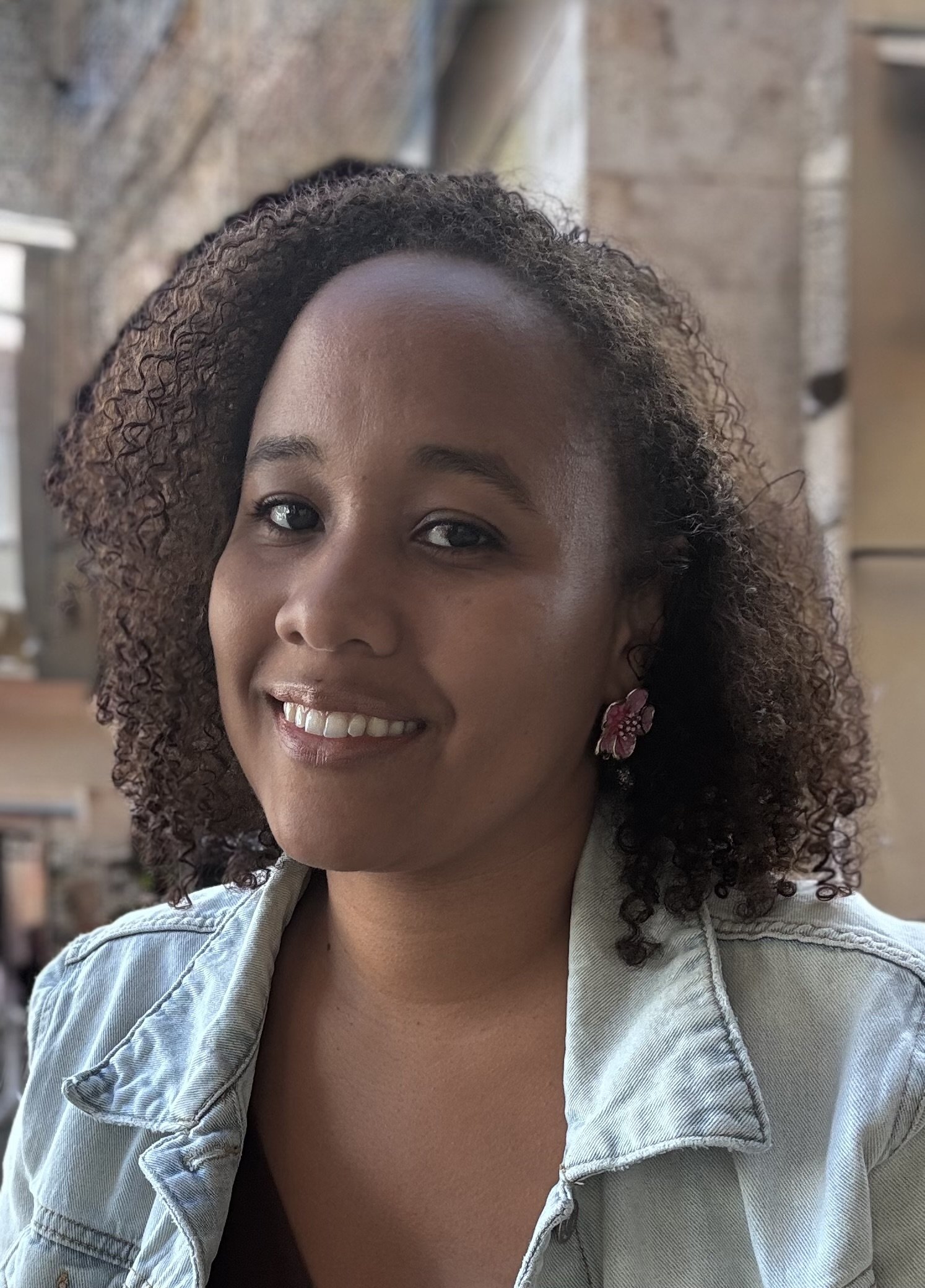 A woman with dark curly hair smiling, wearing a denim jacket and floral earrings, in a cozy indoor setting.