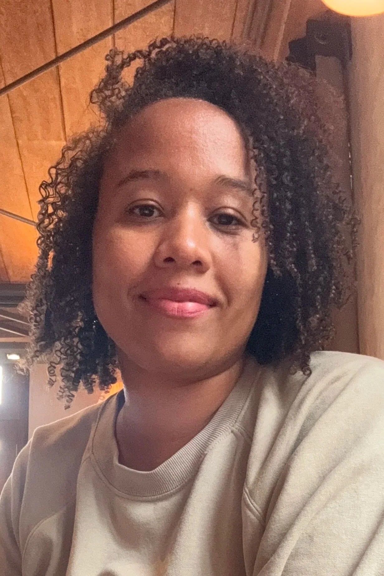 A woman with curly hair smiling in a cozy indoor setting with wooden walls and warm lighting.