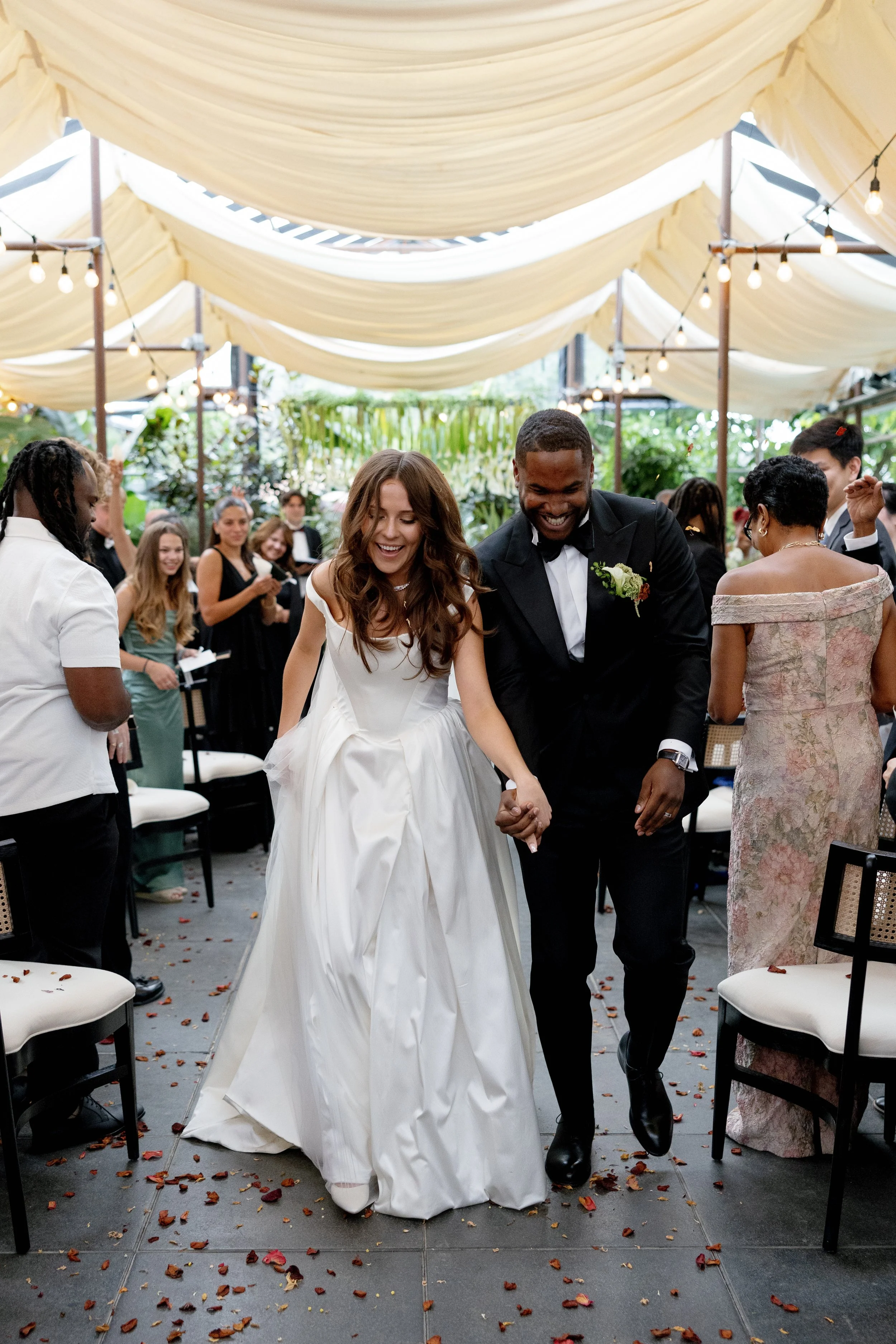 Bride and groom holding hands, smiling and walking down the aisle during their wedding reception, surrounded by guests and decorated with string lights and draped fabric ceiling.
