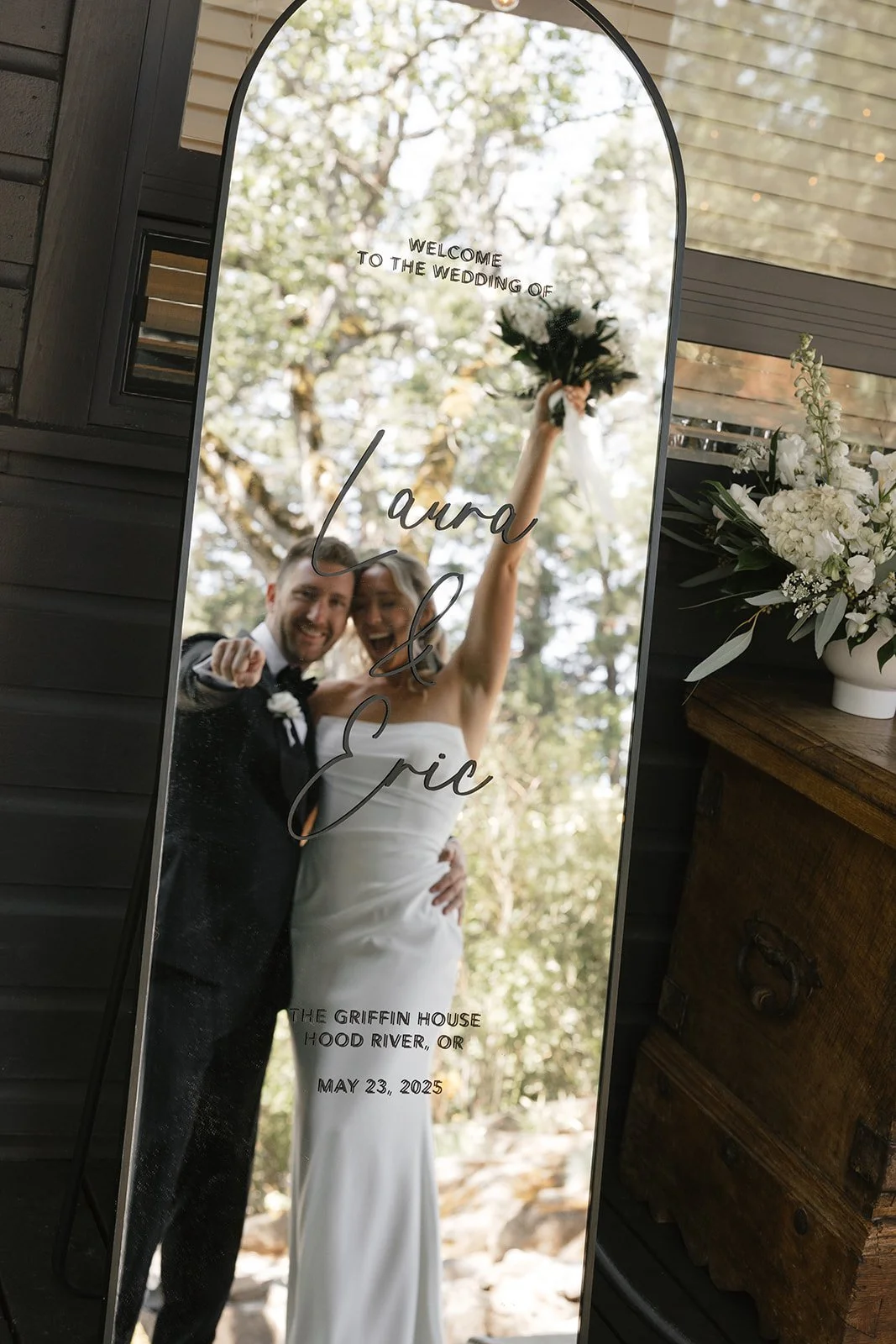 A wedding couple is reflected in a tall, curved mirror with wedding details written on it, including the names Laura and Eric, the date May 23, 2025, and the venue, The Griffin House in Hood River, OR, surrounded by trees and sunlight.