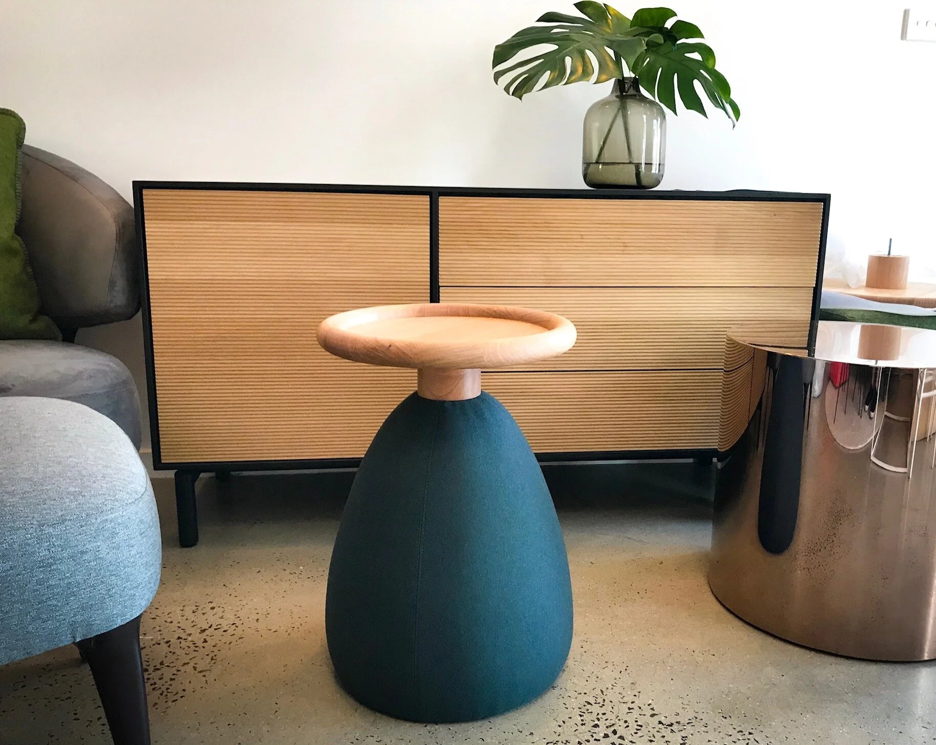 Modern living room with a beige wooden sideboard, a sculptural side table with a wooden top and dark base, a green glass vase with a tropical leaf, a shiny metallic drum table, and a gray upholstered chair.