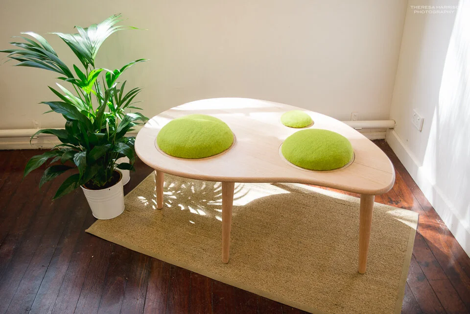Wooden table with green cushions, next to potted plant, in sunlit room on wooden floor and rug.