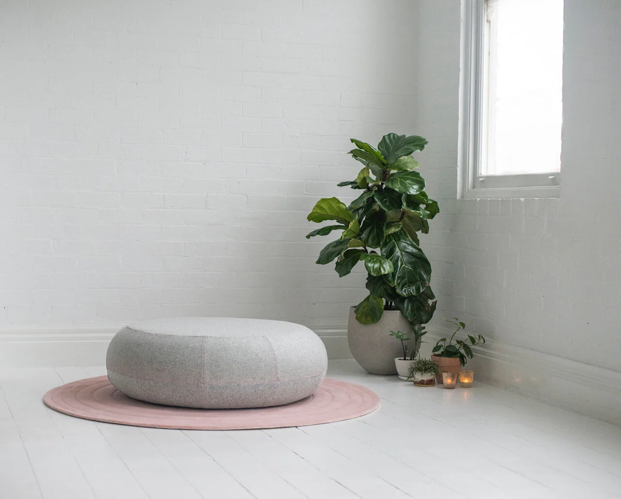 Minimalist room with RICOCHER gray pouf on pink rug, large potted plant, small plants, lit candles, white brick wall.