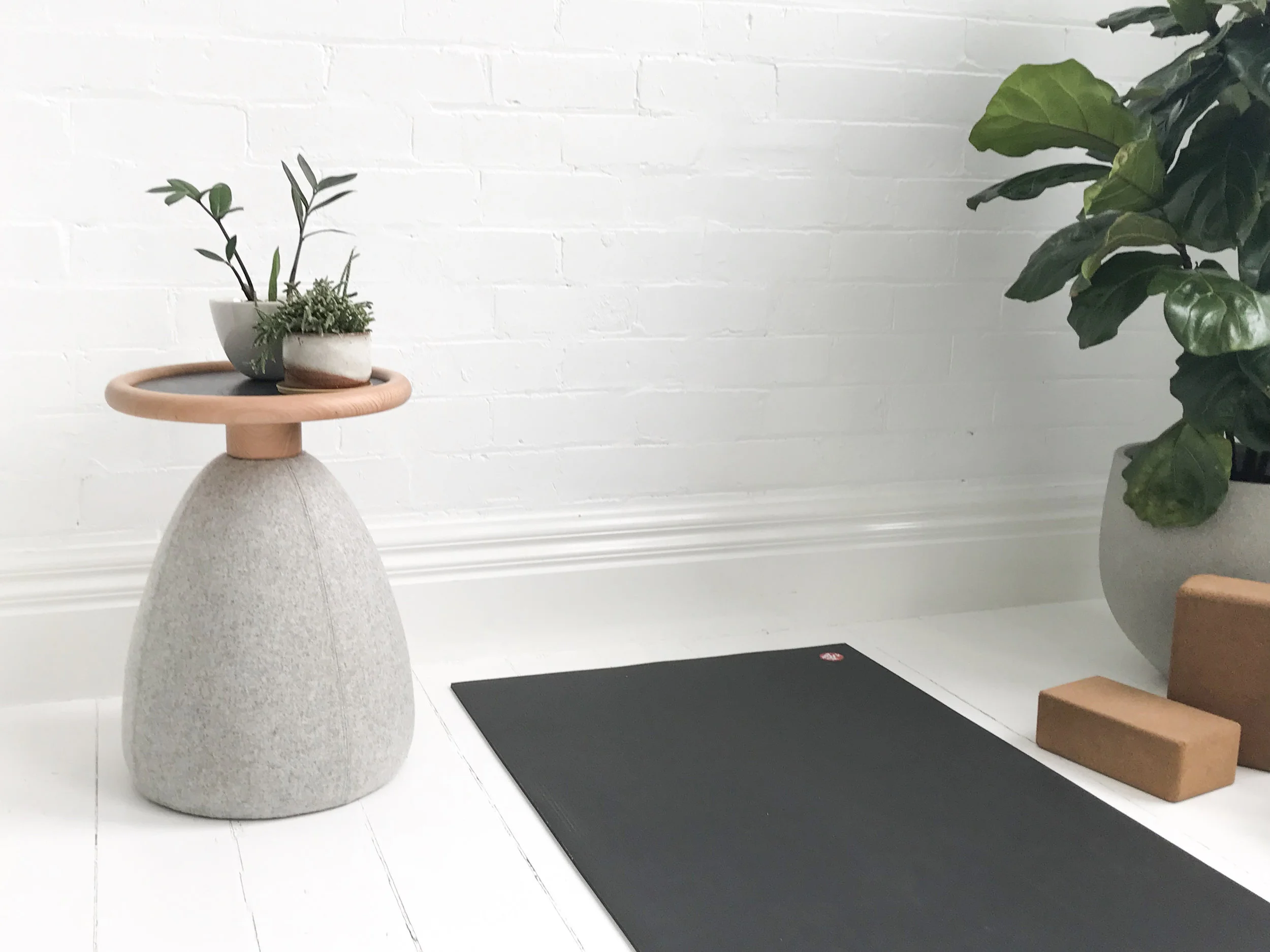 Minimalistic home yoga space with black yoga mat, gray stool with plants, fiddle leaf fig tree, and cork yoga blocks.
