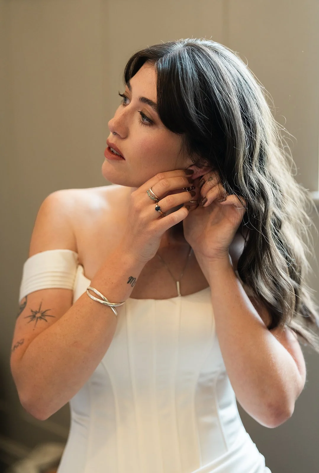 A bride with black and brown hair, wearing a white off-shoulder wedding dress, is putting on earrings while looking to the side. A modern Wedding venue. Bridal Hair & Makeup at Limekiln East sussex