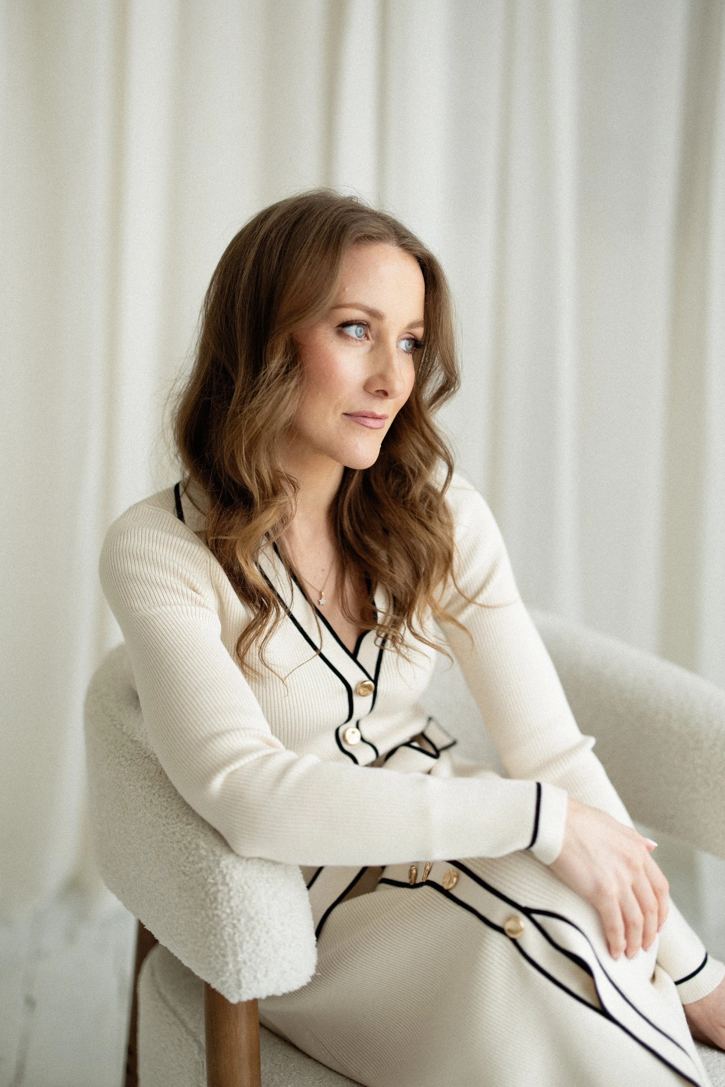 Hair & Makeup artist Lauren Garfitt with light brown, wavy hair and blue eyes sitting in a room with light-colored curtains, wearing a cream-colored ribbed sweater with black trim and gold buttons.