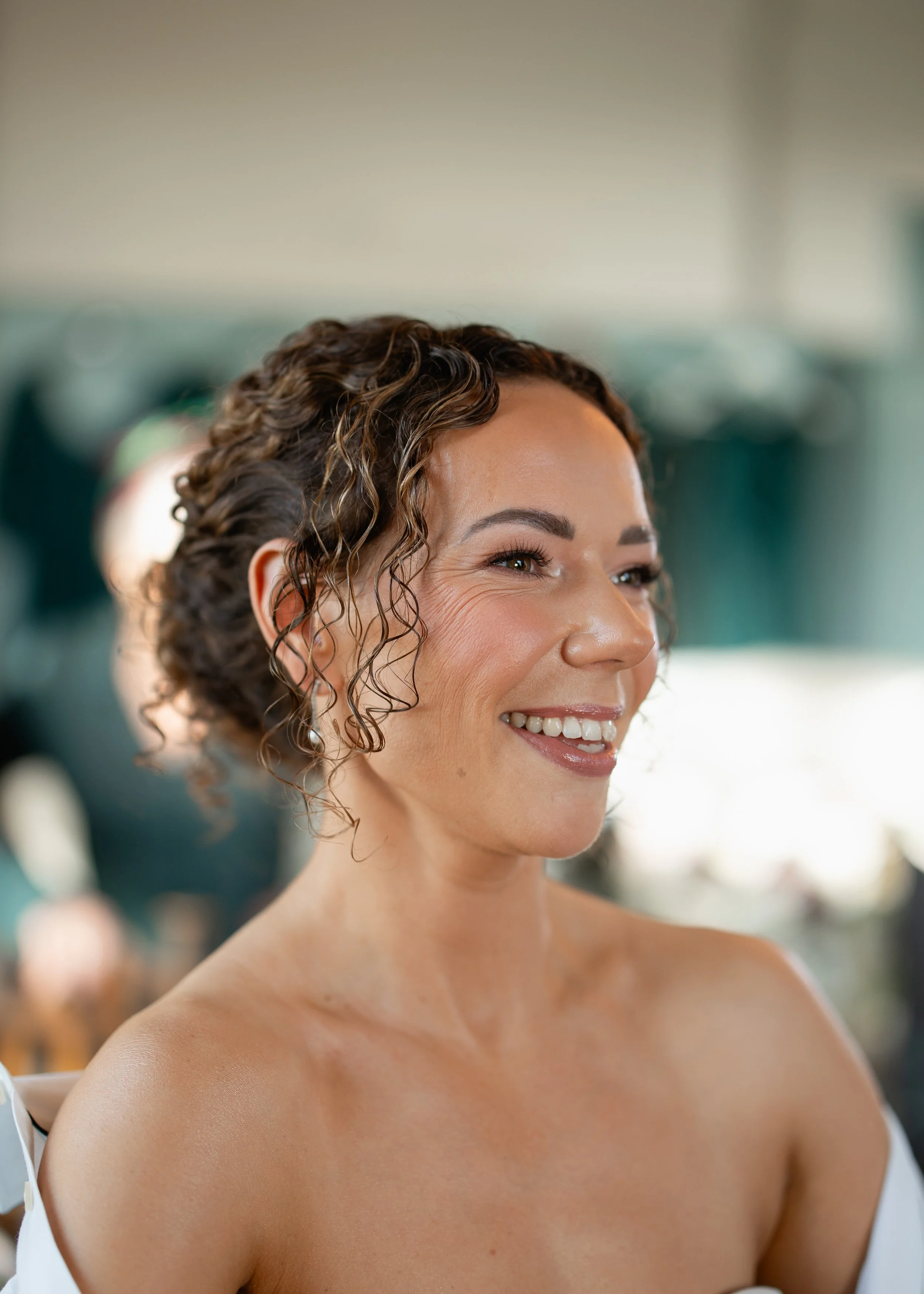 Close-up of a smiling bride with curly brown hair, wearing a white off-shoulder top, with blurred background. Bridal Hair & Makeup at The Beacon