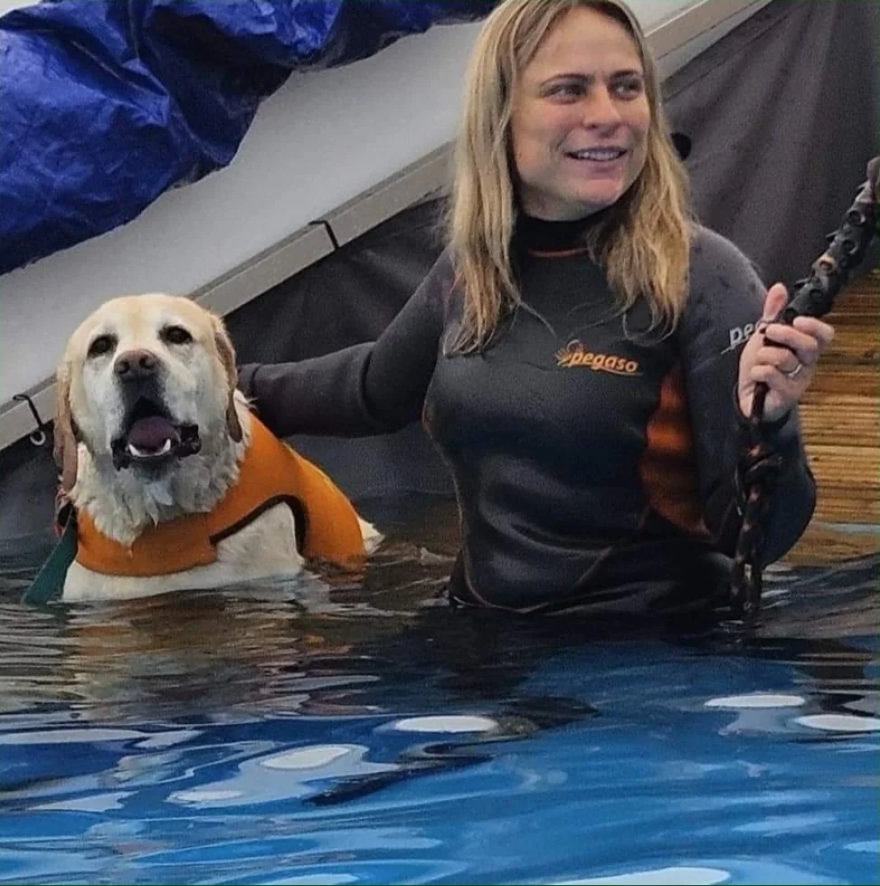 A woman and a dog in a body of water, possibly during a water rescue training exercise. The woman is holding a rescue tool, and the dog is wearing a life jacket.