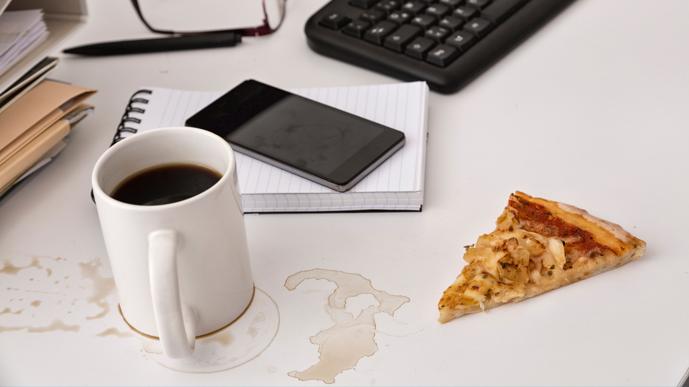An office workers' dirty desk that has a notepad, cellphone, pizza not on a plate, a dirty coffee cup and coffee stains on the desk surface