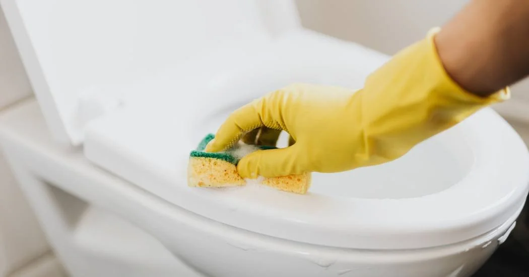 a person wearing yellow rubber gloves scrubs a toilet seat with a sponge