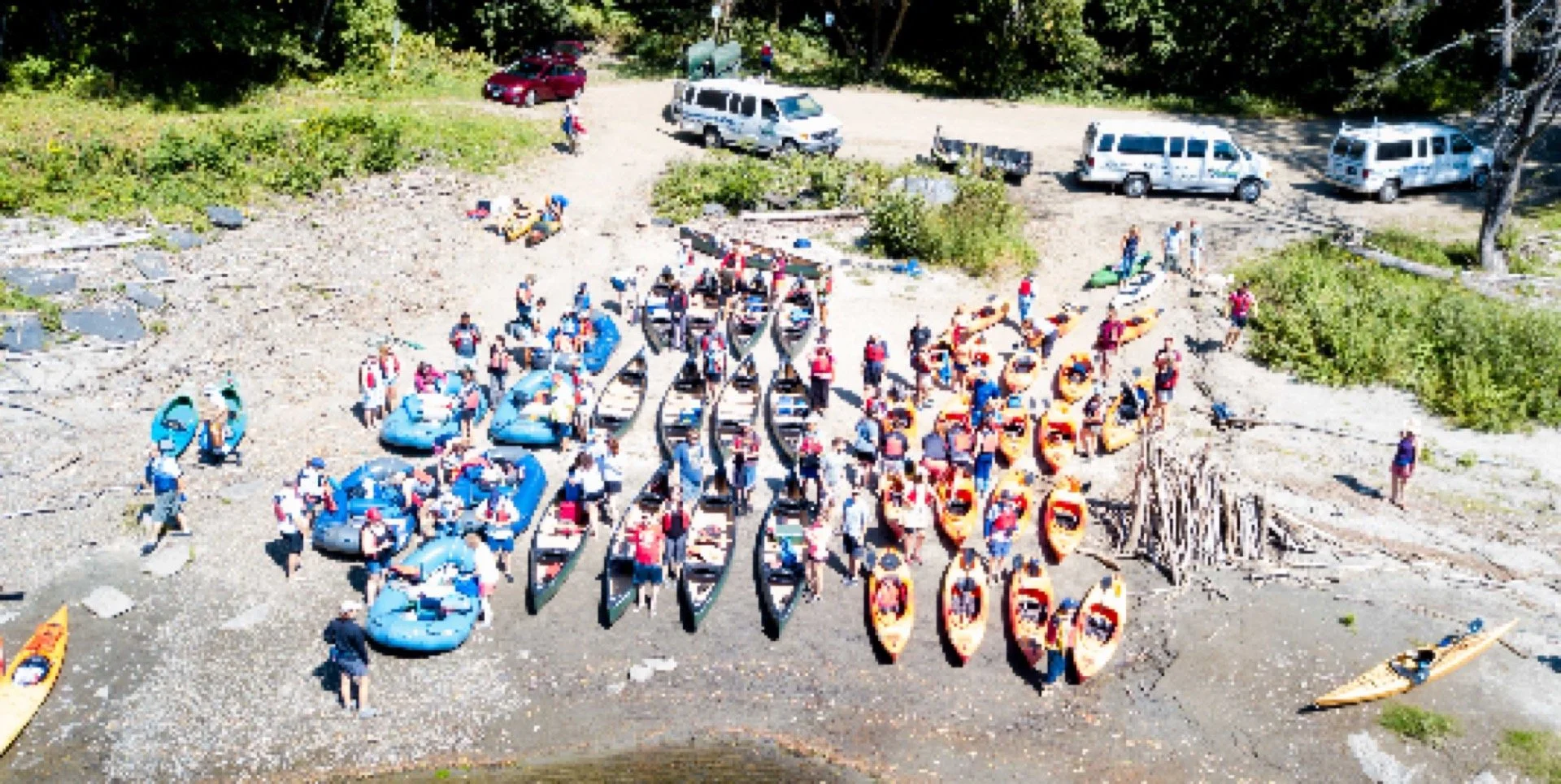 People preparing for kayaking on a rocky beach, with kayaks and paddleboards lined up, and vehicles parked in the background.