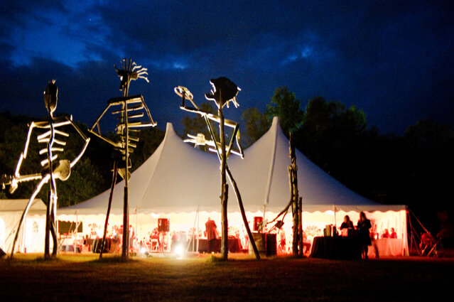 Outdoor event tent illuminated at night, decorated with tall skeleton-like sculptures with lights, and trees in the background.