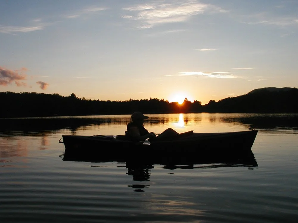 A person sitting in a kayak on a calm lake at sunset, with the sun near the horizon and a silhouette of trees and hills in the distance.