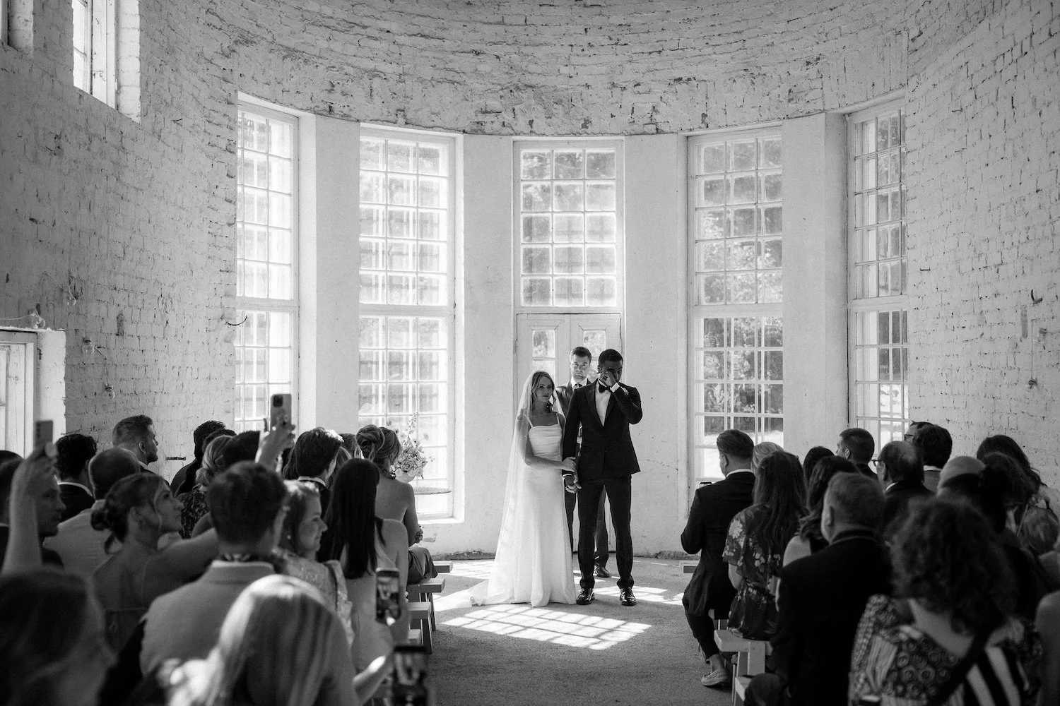 Hääseremonia, missä morsiuspari ja vihkijä seisovat julkisivun edessä, kahdeksan taustalle järjestäytynyttä suurikokoista ikkunaa ja valkoiset seinät taustalla, häävieraat istuvat ja seuraavat tilannetta, osa ottaa kuvia.