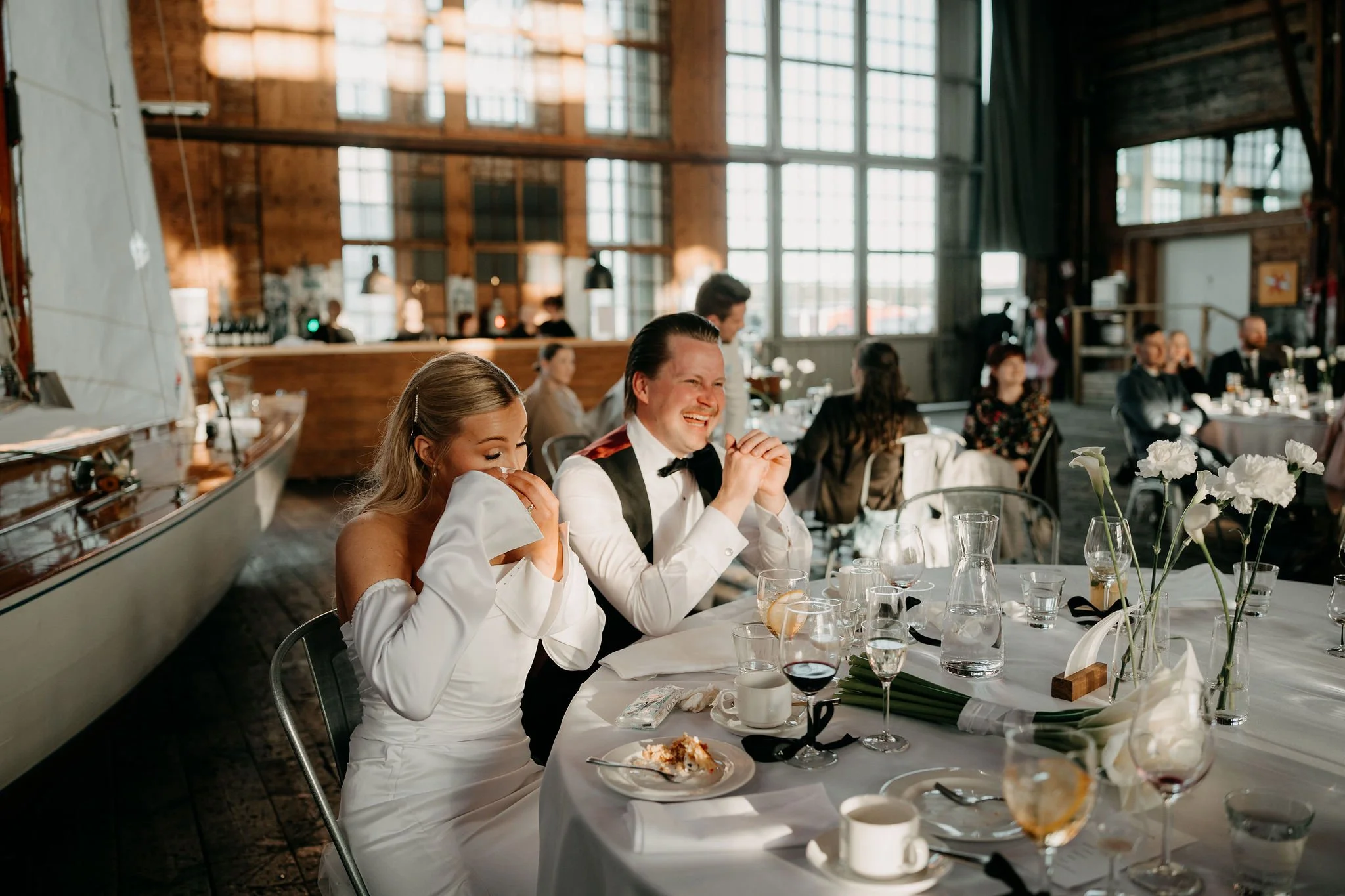 Hääpari istuu vierasjoukkojen keskellä juhlavaatteissa juhlasalissa, nauravat ja nauttivat: