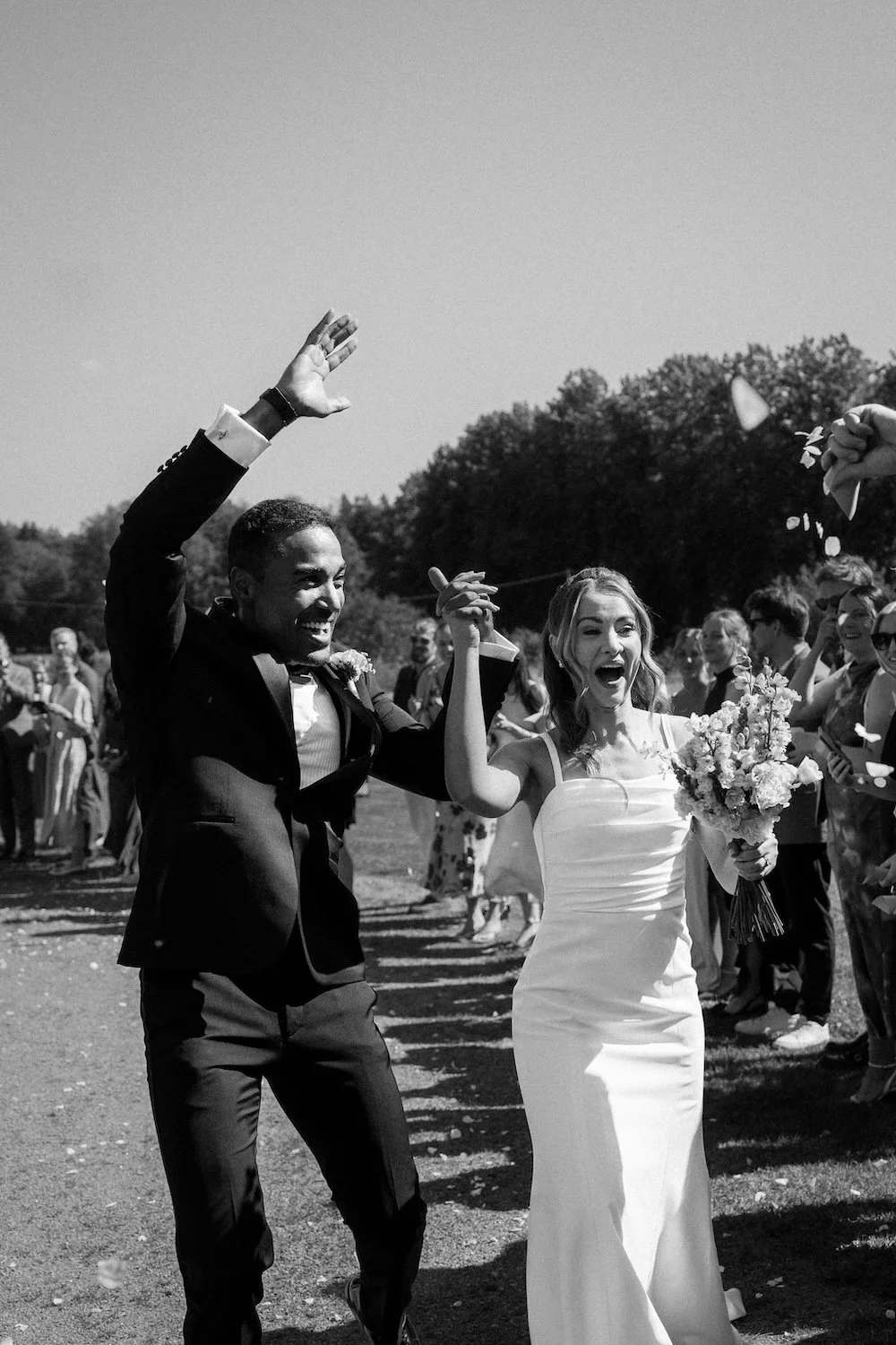 Värillisenä kuva pariskunnasta hääasussa iloisesti juhlimassa ulkona, kädet nostettuina iloisina, hymyilevät ja kukka-asetelma