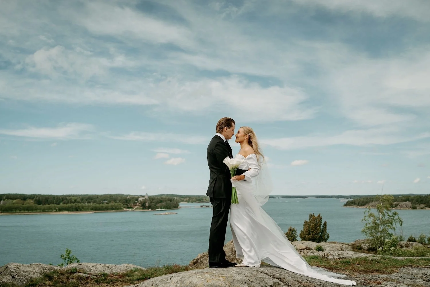 Hääpari seisoo rannalla, mies ja nainen katselevat toisiaan, nainen pitää kukkakimppua, taustalla järvimaisema ja sininen taivas.