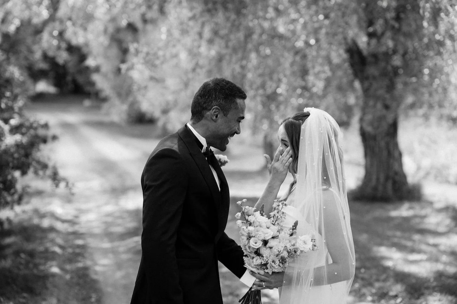 Hääpari, mies ja nainen, hymyilevät ja katsovat toisiaan, nainen pitää kukkakimppua, taustalla puut ja polku