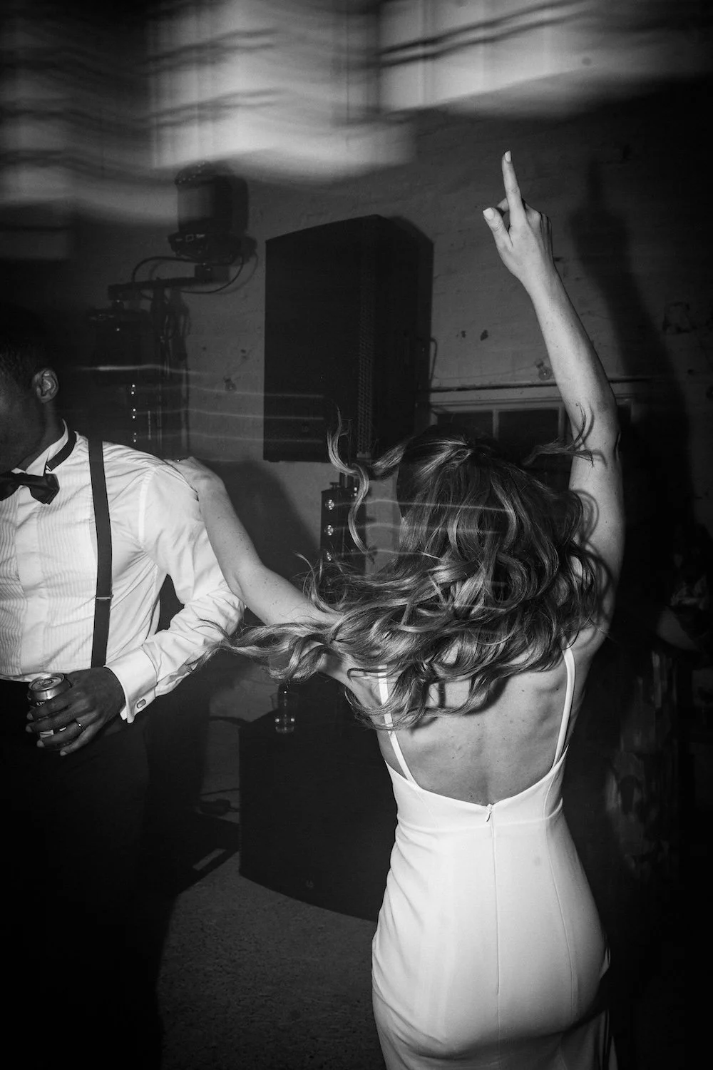 Mustavalkokuva ihmisistä tanssimassa juhlatilassa, nainen heiluttaa hiuksiaan ja tekee käsiliikkeen, mies pitää juomapulloa, taustalla kaiutin ja valoja.