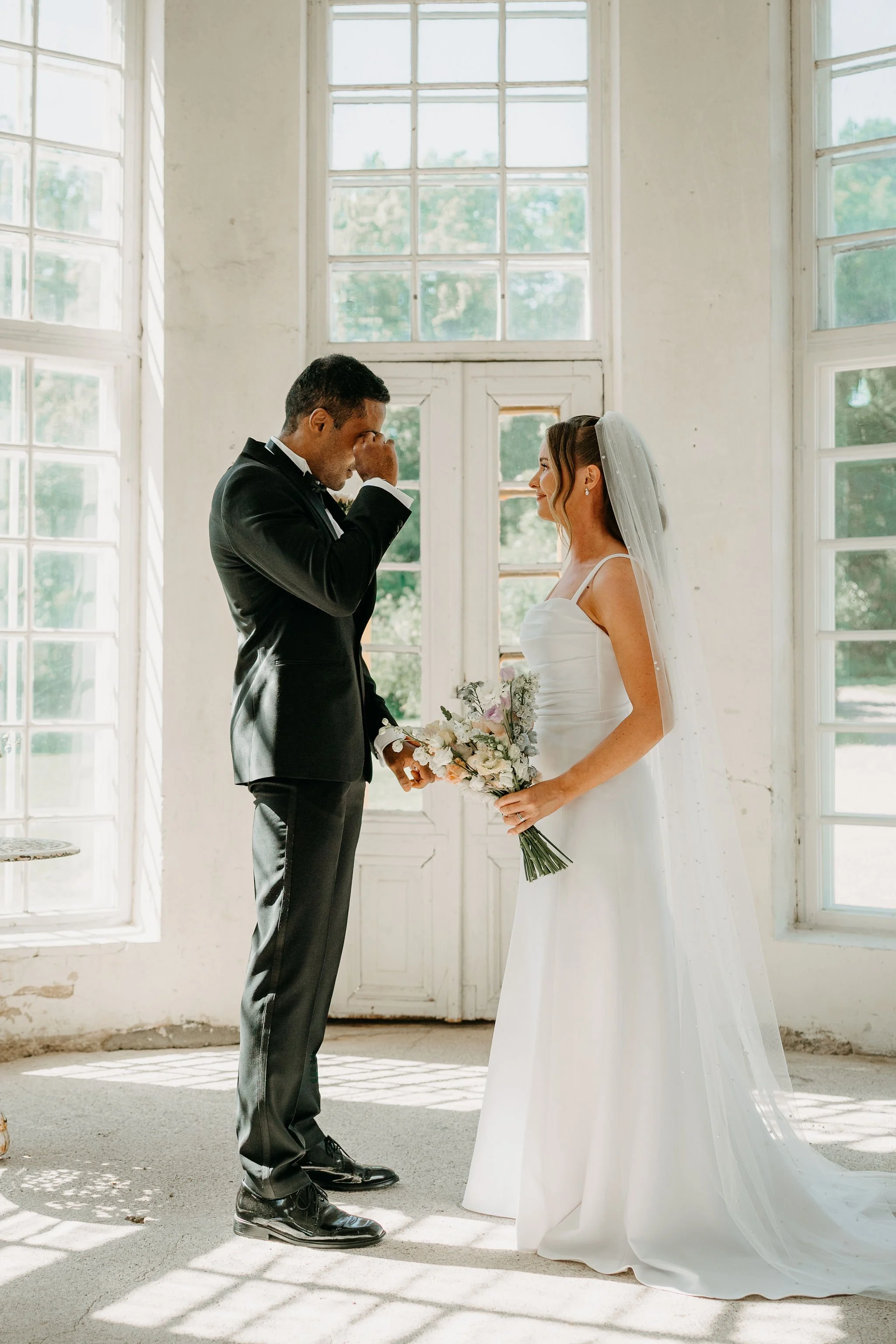 Häät: Mies ja nainen seisovat sisällä valossa, nainen pukeutuu valkoiseen pukuun ja mies pukuun, nainen pitää kukkakimppua, nainen hymyilee ja mies itkee