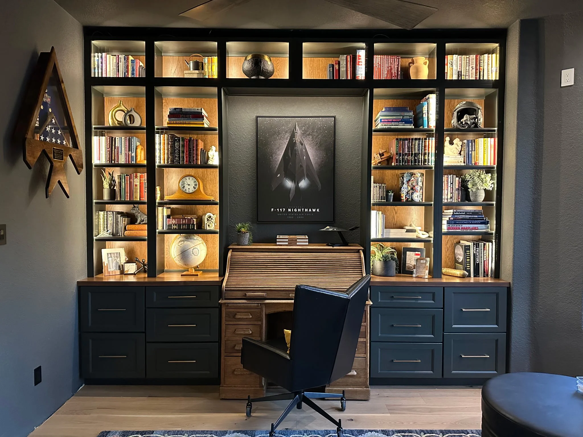 A home office with a built-in bookshelf, a black office chair, and a wooden writing desk. The bookshelf contains books, decorative items, and framed photos. A central framed poster of an aircraft is on the wall behind the desk. There is a cabinet wit