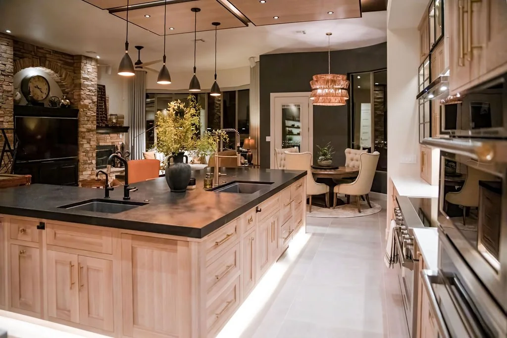 Modern open-concept kitchen with light wood cabinetry, black countertops, and pendant lighting, leading to a dining area with a round table and upholstered chairs, and a living room with a stone fireplace and large windows.