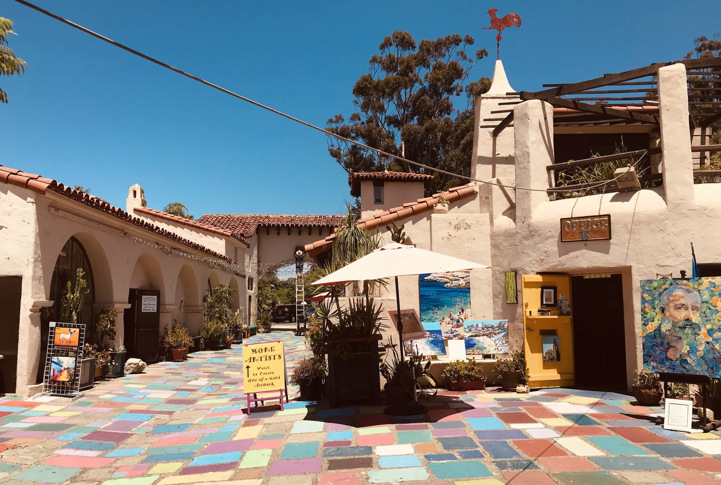 An outdoor art gallery with colorful painted tiles on the ground, displaying paintings and art pieces along a white stucco building with a red tile roof under a clear blue sky.