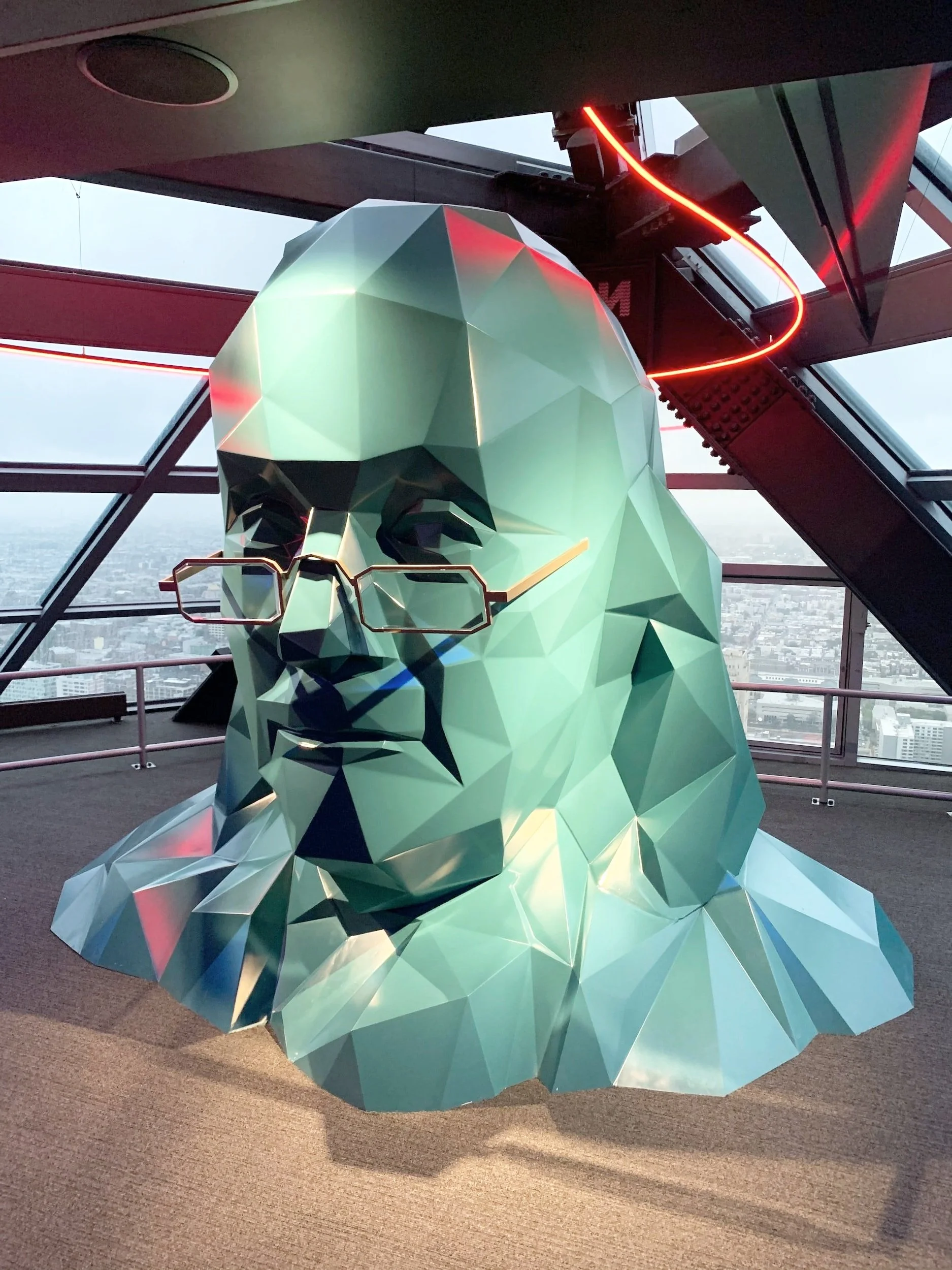 A geometric sculpture of a human bust with glasses, placed inside a glass-walled high-rise building.
