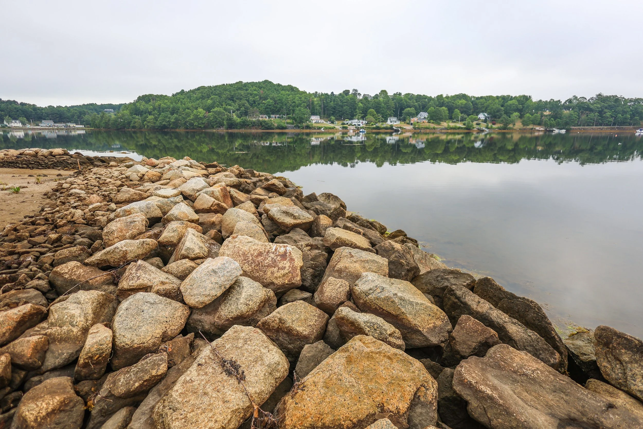Rocky shoreline with a calm lake reflecting a green hill and houses.