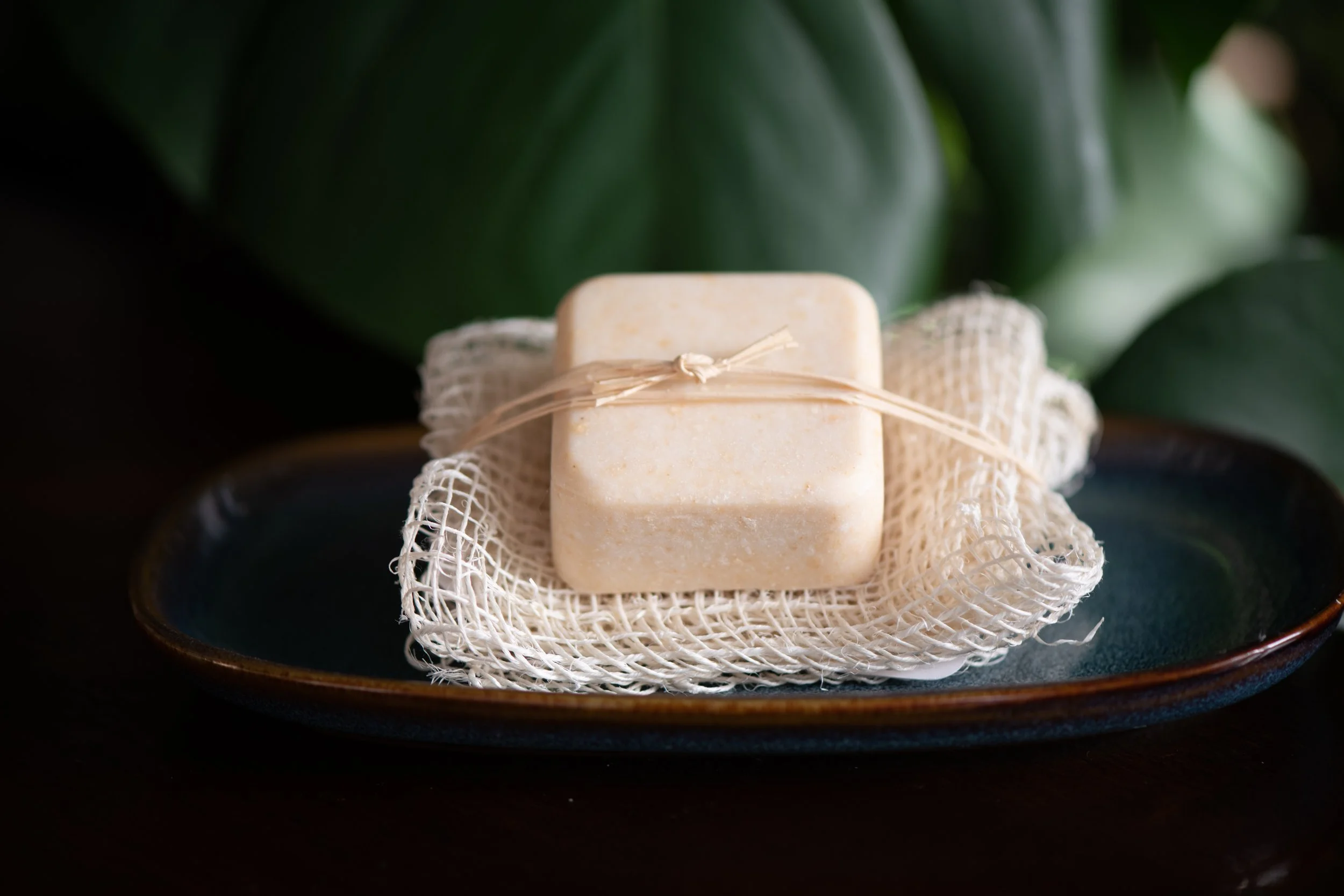 Wayward Bison Golden Glow Soap with Ayate Fiber Cloth. Pale orange, two ounce bar of soap on a folded agave fiber washcloth sitting on a deep blue ceramic dish with a plant in the background.
