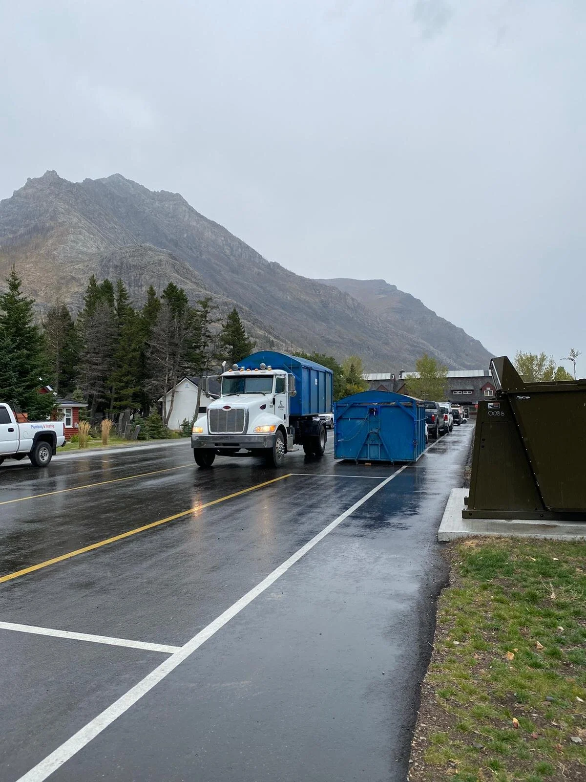 View of a rainy street with a white truck, a blue waste bin, and parked cars against a backdrop of mountains and trees.