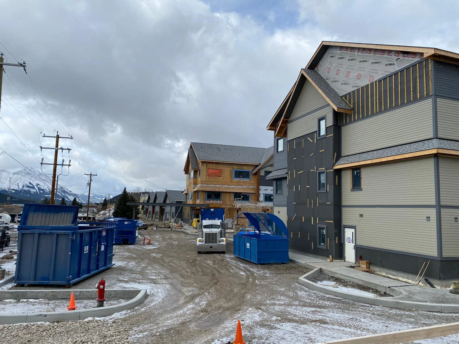 Under construction multi-story residential buildings with construction bins and equipment on muddy ground, snow, and cloudy sky, with mountains in the background.