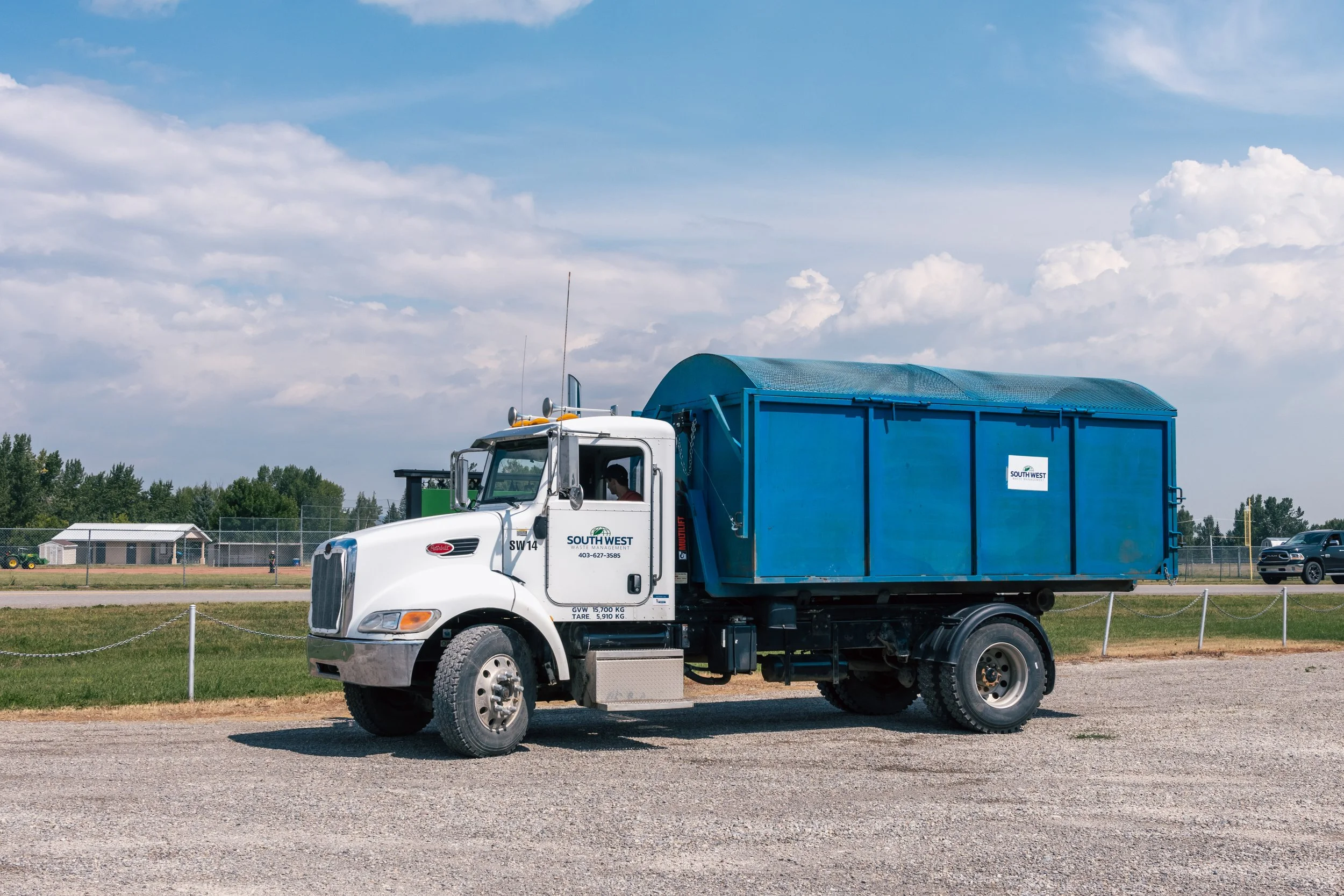 A white and blue waste management truck parked on gravel, with a running track, grass field, and school buildings in the background under a partly cloudy sky.