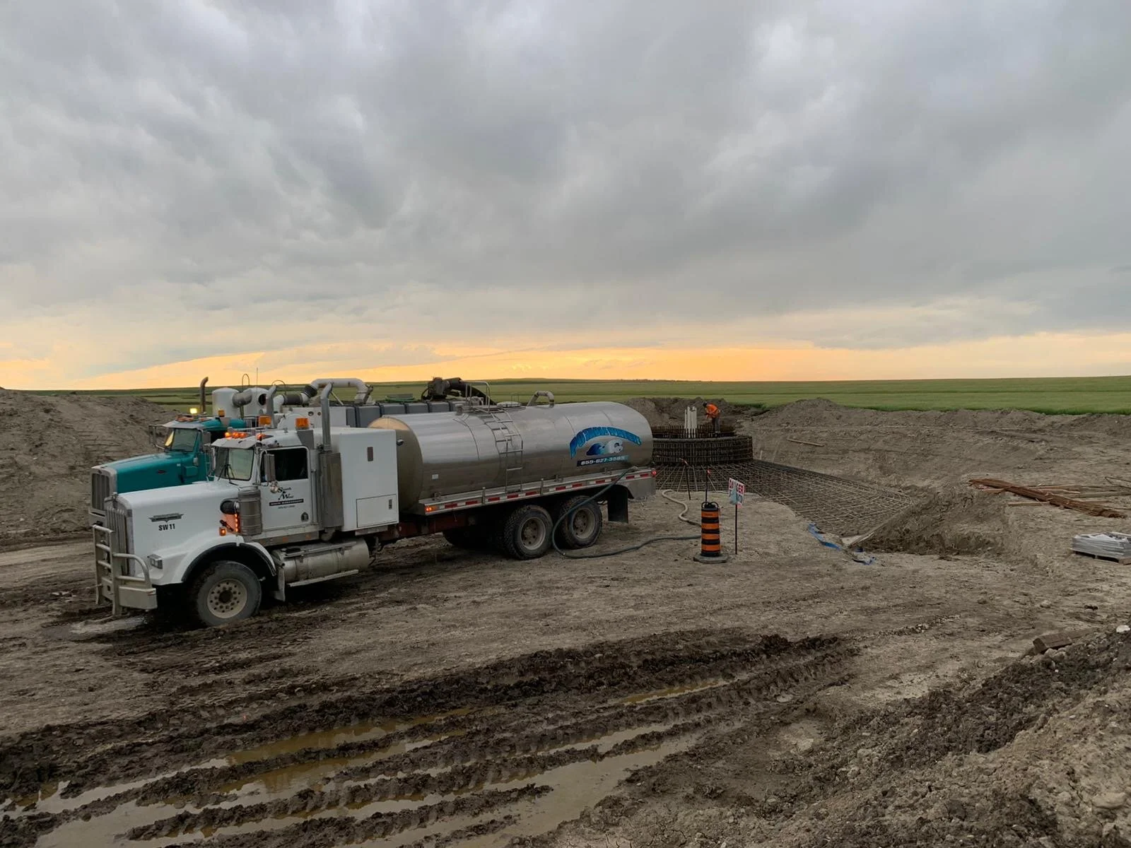 Construction site with a large tanker truck parked on muddy soil, with workers and metal rebar grid in background, under cloudy sky at sunset.