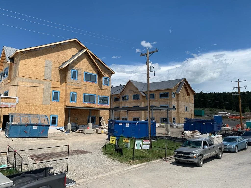 Construction site with partially built multi-story houses with wooden framing, blue window frames, construction dumpsters, parked vehicles, and utility poles under a partly cloudy sky.