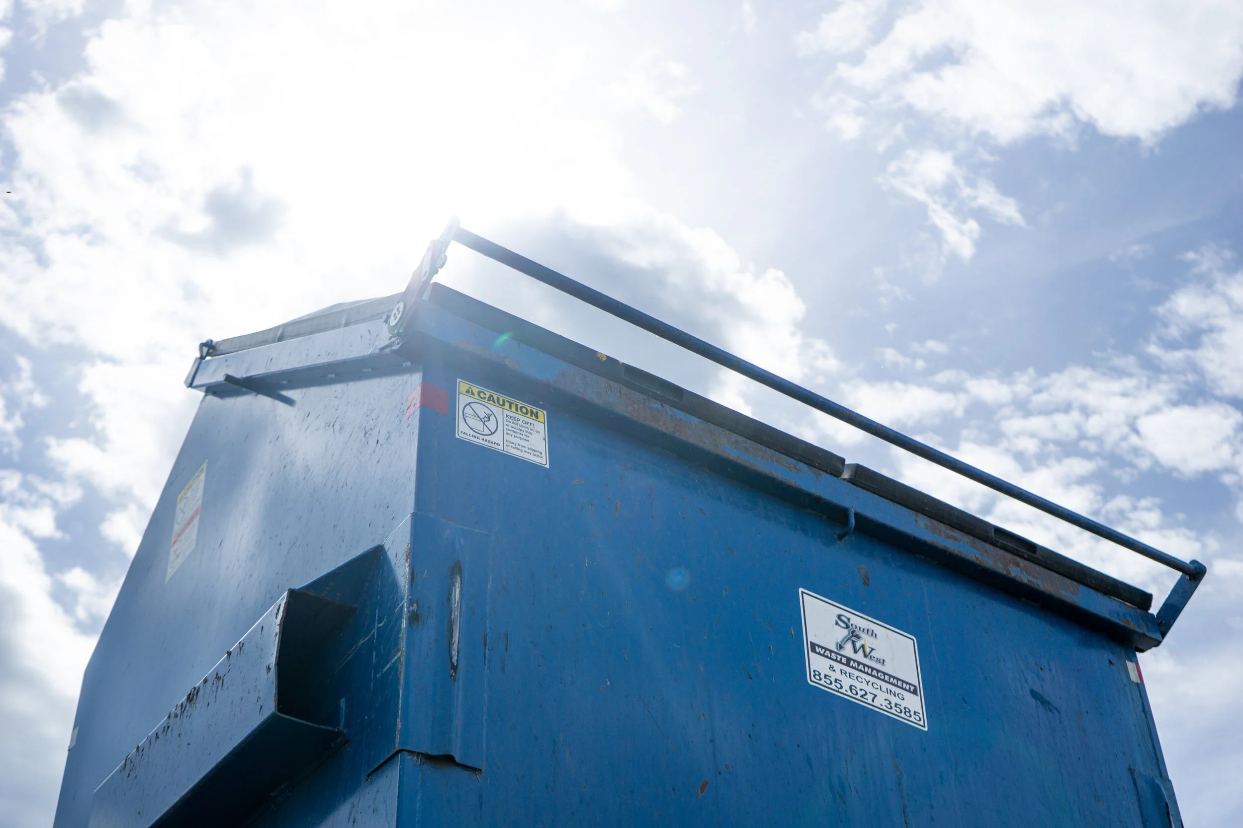 Blue industrial waste or recycling dumpster with caution and contact stickers, set against a partly cloudy sky with sunlight glare.