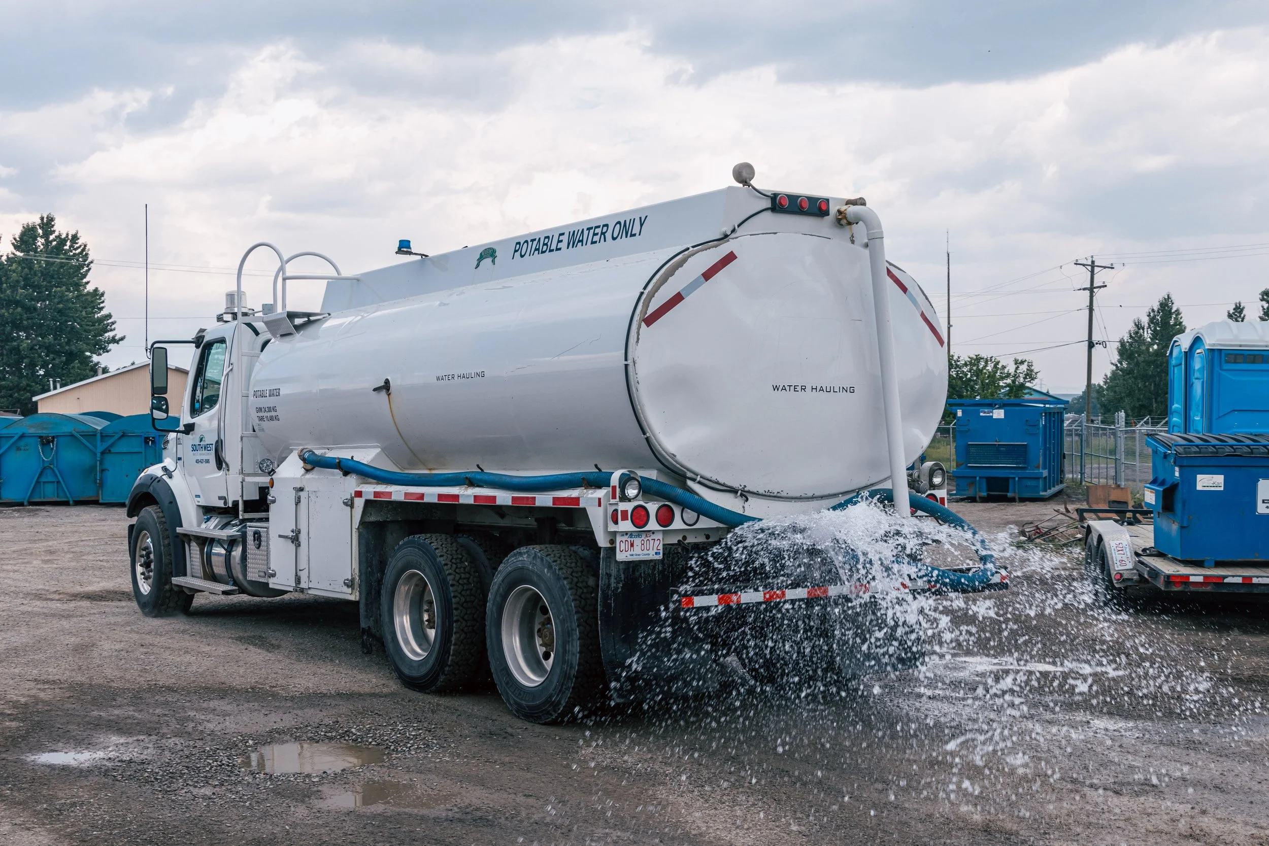 White water tanker truck spraying water on the ground, with blue containers and a cloudy sky in the background.