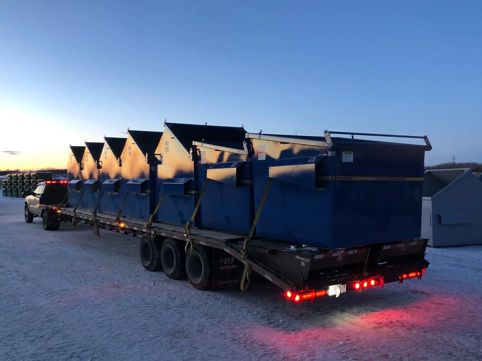 Blue industrial dumpsters on a flatbed trailer truck parked on a snowy ground at dusk.