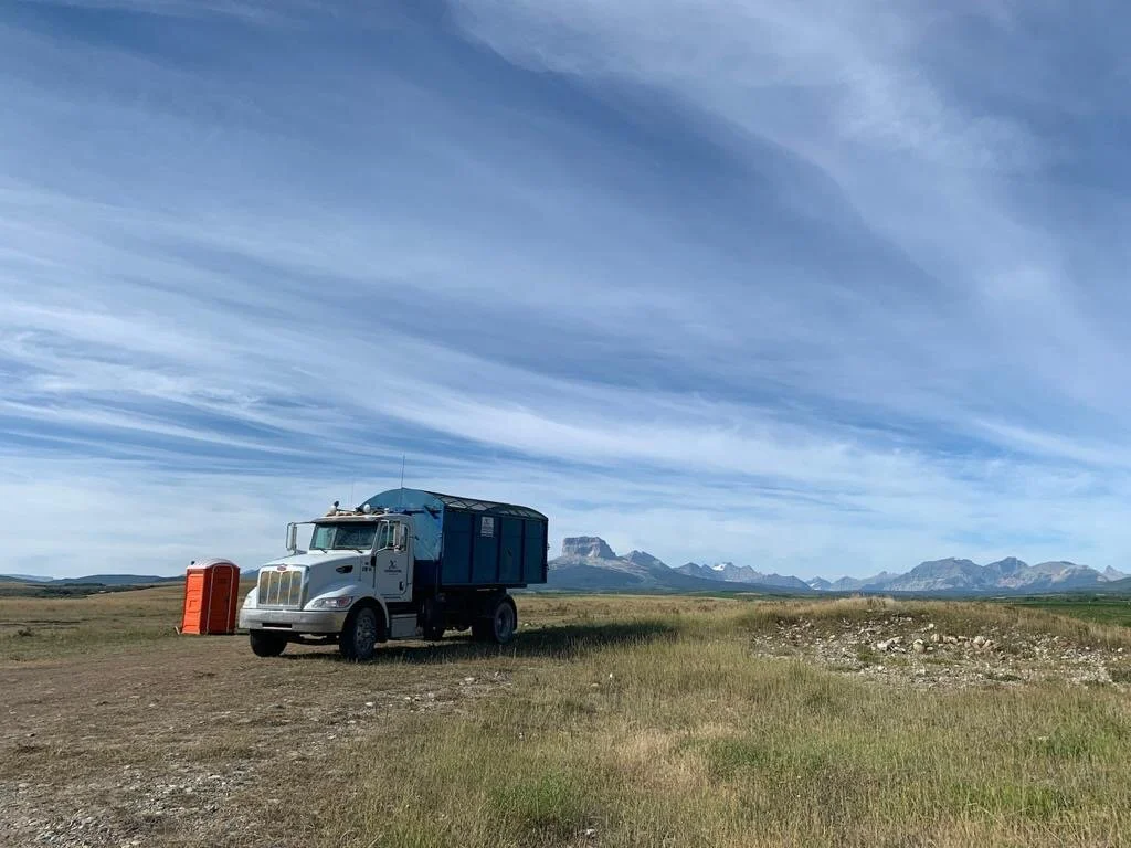 A large truck parked on a grassy plain with mountains in the background, a new orange portable toilet nearby, under a blue sky with wispy clouds.