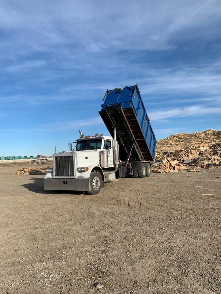 A white dump truck with its blue container raised, dumping debris or wood chips on a construction site or landfill, with a clear blue sky overhead.