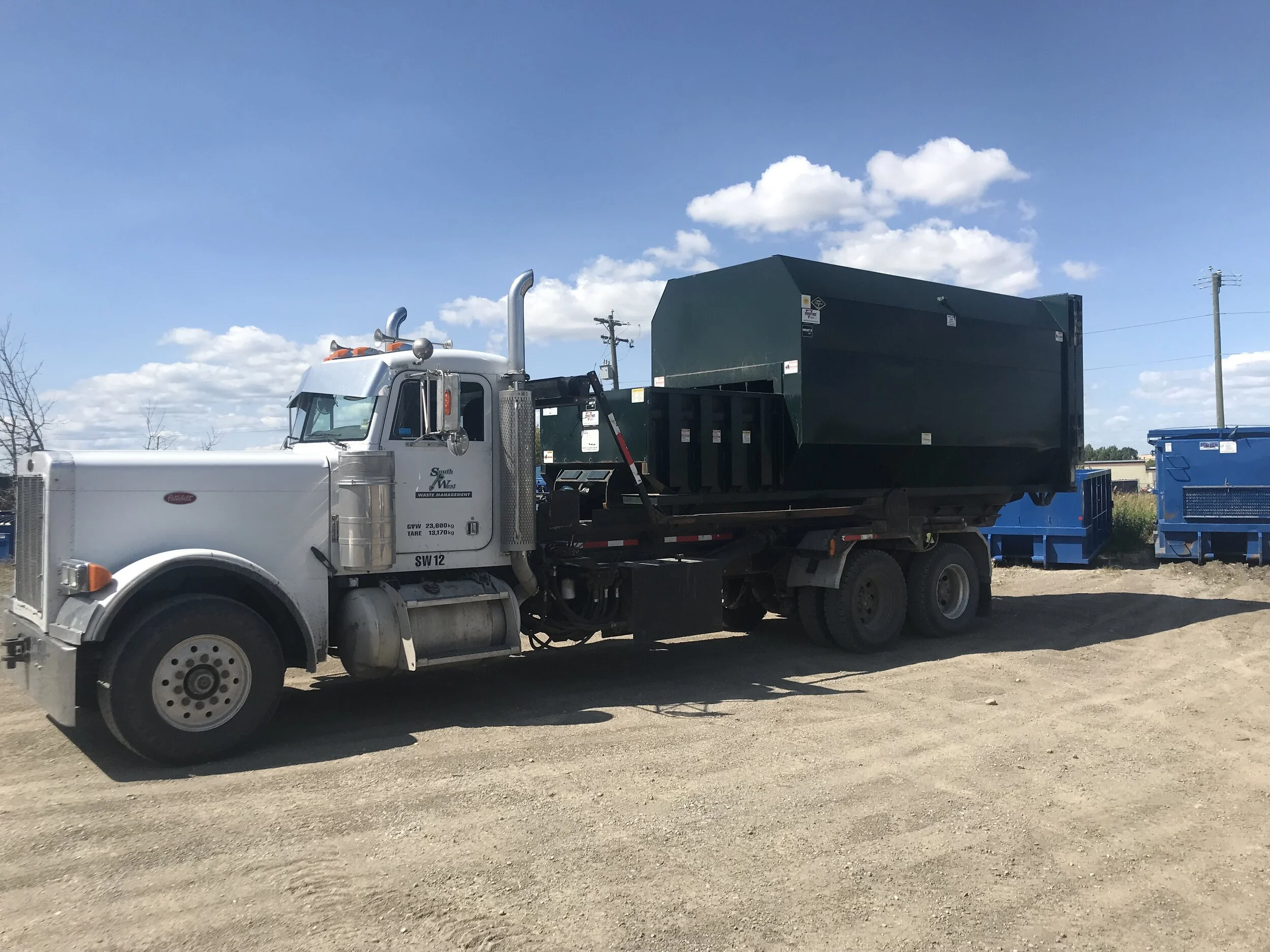 A large white and black garbage truck parked on a dirt lot, with blue dumpsters in the background under a partly cloudy sky.