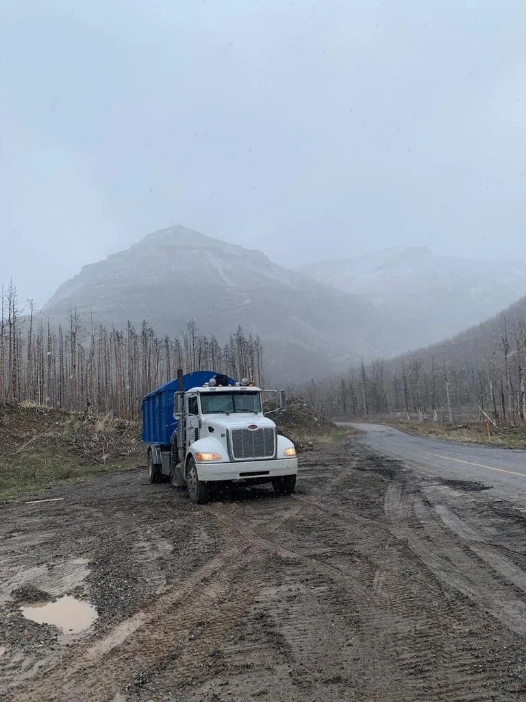 White and blue dump truck on a muddy mountain road with trees and mountains in the background.